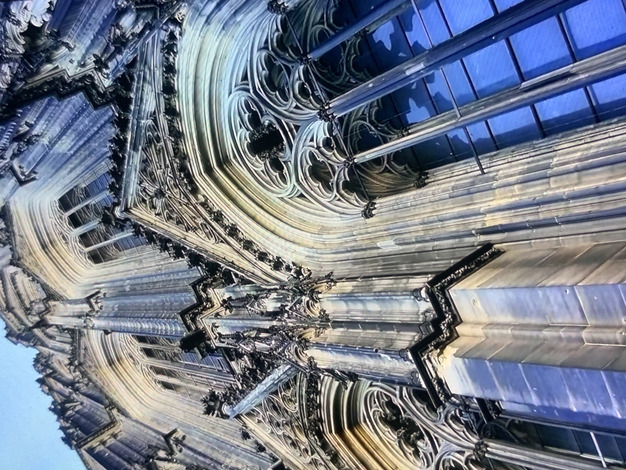 The image displays a low-angle shot of the exterior of a grand Gothic cathedral. The architecture features intricate stonework, pointed arches, and ornate decorative elements, including tracery in the windows. Tall, narrow windows with a vibrant blue tint are visible, reflecting the sky. The stonework is a mix of grey and lighter, weathered tones. The scene is devoid of people or any other discernible objects. The sky above is a clear, light blue. The provided context of Stavropol, Russia, does not align with the architectural style depicted, which is characteristic of Western European Gothic cathedrals.