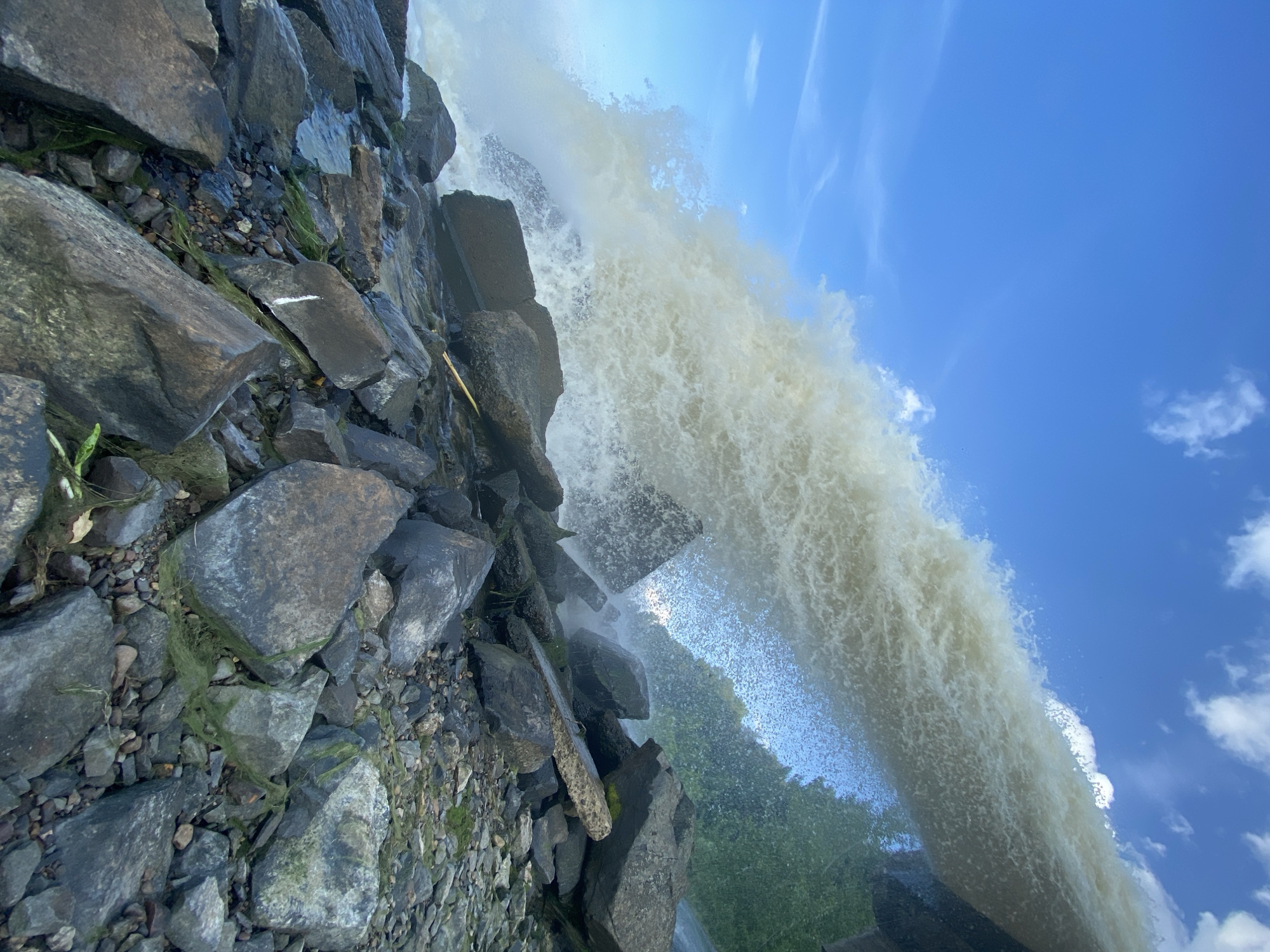 The image depicts a powerful waterfall cascading over rocks under a bright blue sky with scattered white clouds. The water is a turbulent, foamy white, suggesting a significant flow. The foreground is dominated by a rocky shore, with large, dark grey stones of various sizes. Some of these rocks are covered in patches of green algae or moss, indicating a damp environment. A thin stream of water flows between some of the rocks in the lower left of the frame. In the background, lush green foliage is visible on the right side, suggesting trees or vegetation bordering the waterfall. The overall impression is one of natural power and a dynamic, watery environment. The location is identified as the city of Chernushka, Russia, implying this is a natural or man-made waterfall within that geographical area. There are no people or animals visible in the image.
