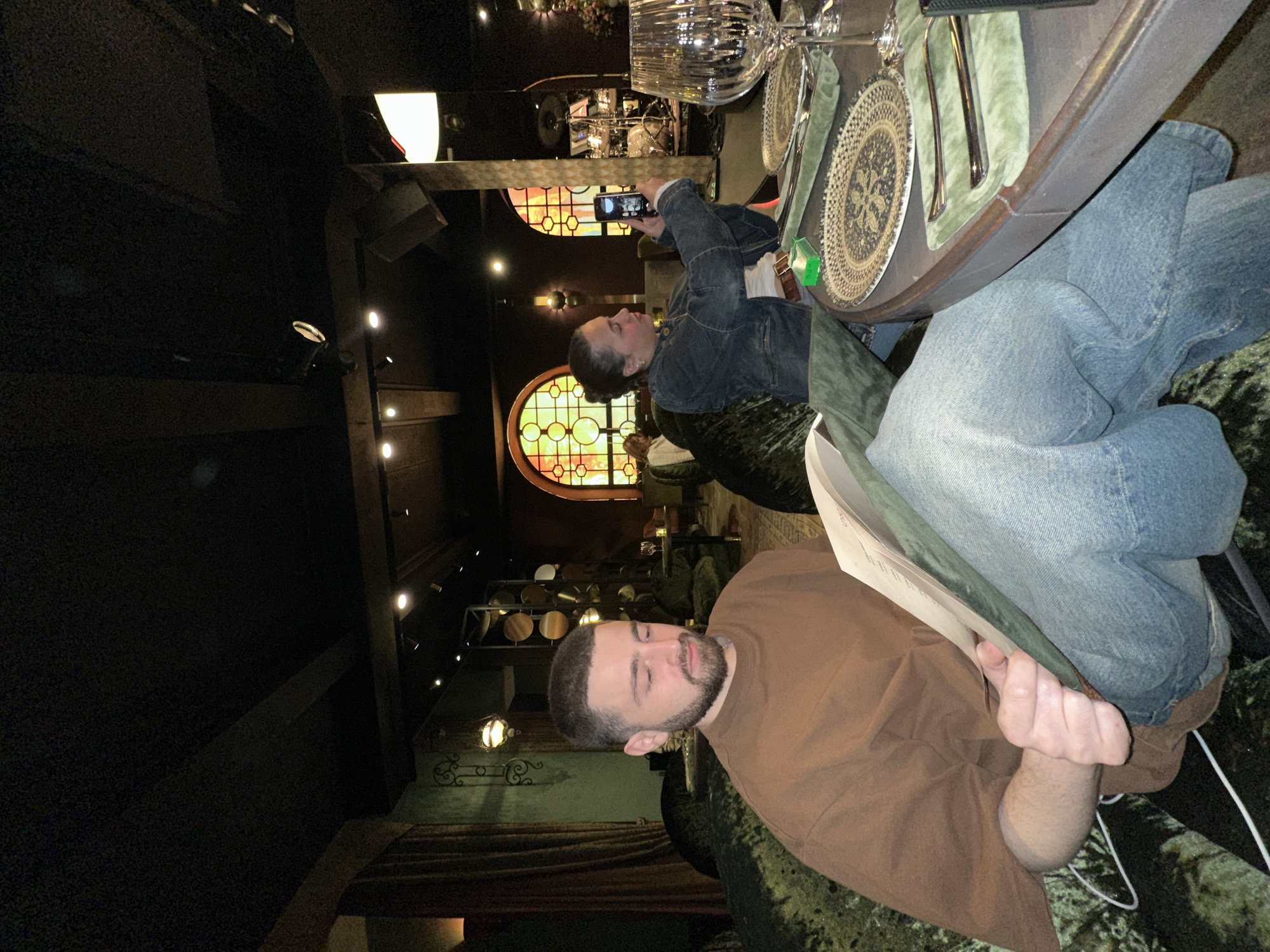 In a dimly lit restaurant, possibly in Yerevan, Armenia, two individuals sit at a table set with ornate chargers and silverware. One person, wearing a denim jacket and jeans, holds up a smartphone to take a photo, presumably of the setting. The other person, in a brown shirt, looks down at a menu. Arched stained-glass windows are visible in the background. The environment includes plush velvet seating and decorative lighting.