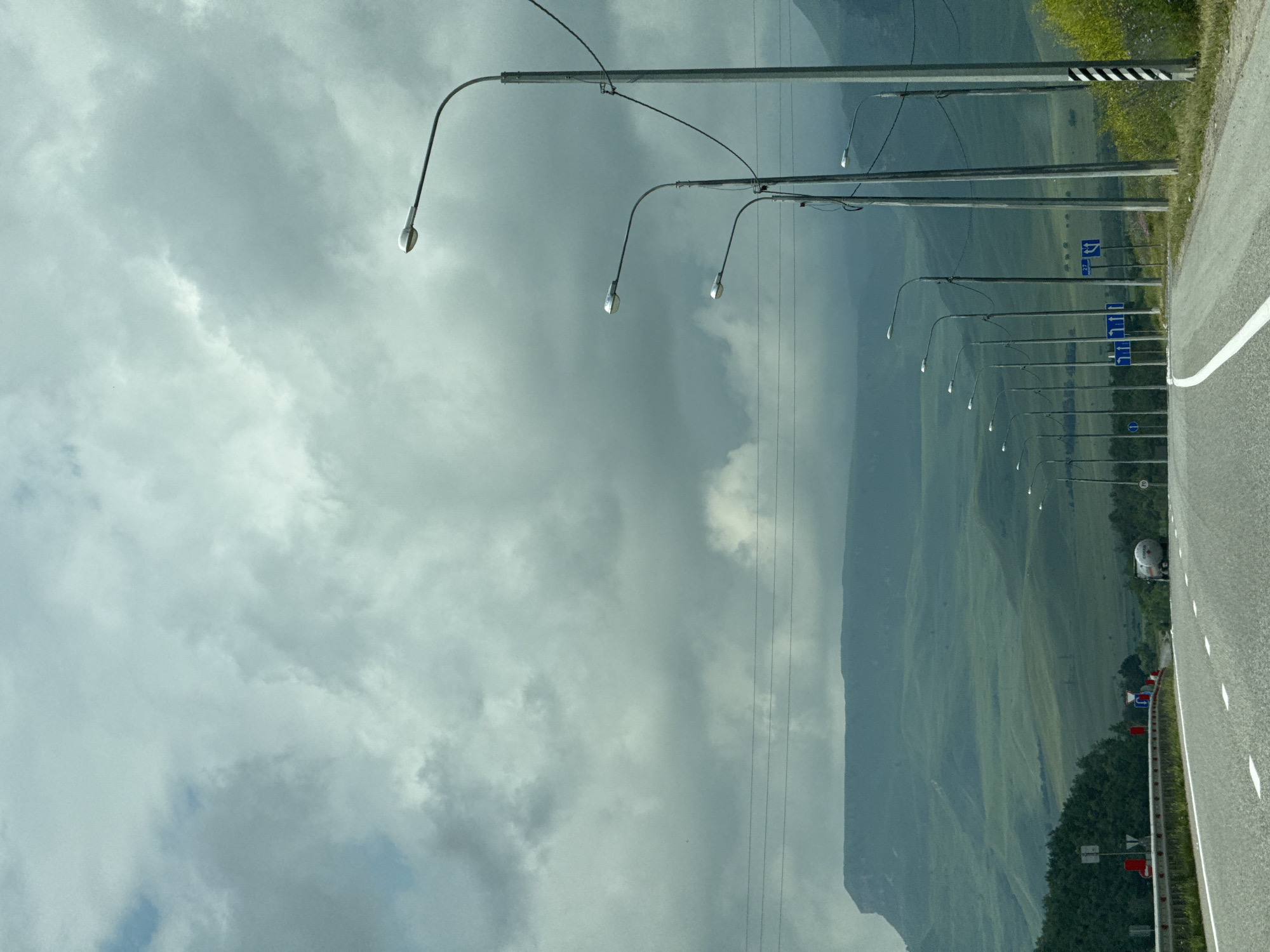 The image depicts a road scene on a cloudy day, likely on the outskirts of Kubina, Russia, given the presence of a road sign indicating "27" which could represent a route number or distance. The road, paved and divided by white lines, curves to the right and is flanked by a metal guardrail on the left. On the right side of the road, a series of streetlights are visible, extending into the distance. Beyond the road and streetlights, rolling green hills form the backdrop, with a prominent flat-topped mountain range in the far distance. Trees line the left side of the road, and a white tanker truck is visible further down the road. The sky is heavily overcast with gray clouds.