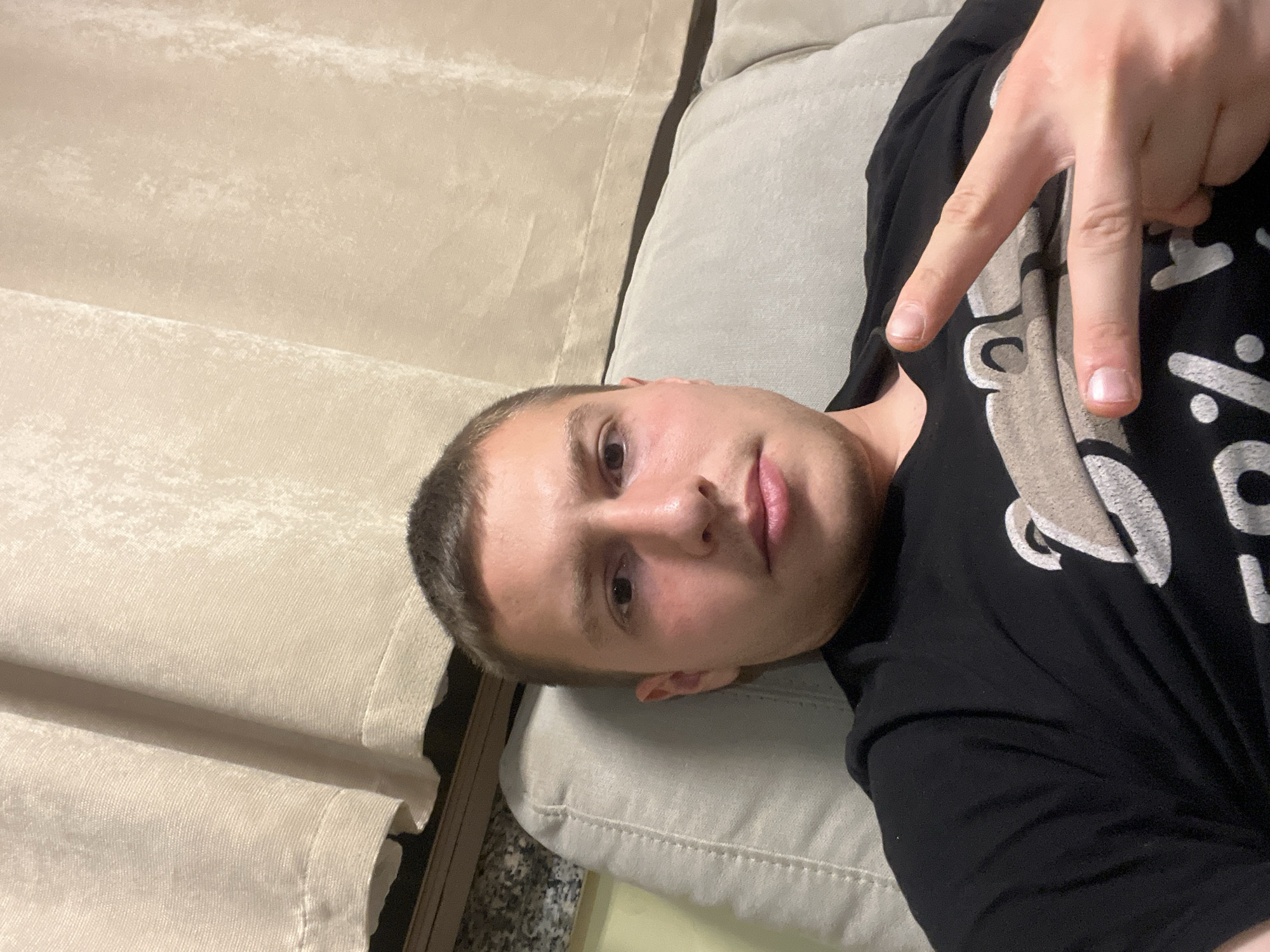 A man is lying on a beige sofa, wearing a black t-shirt and making a peace sign with his right hand. There are beige curtains in the background. The location cannot be determined from the image.