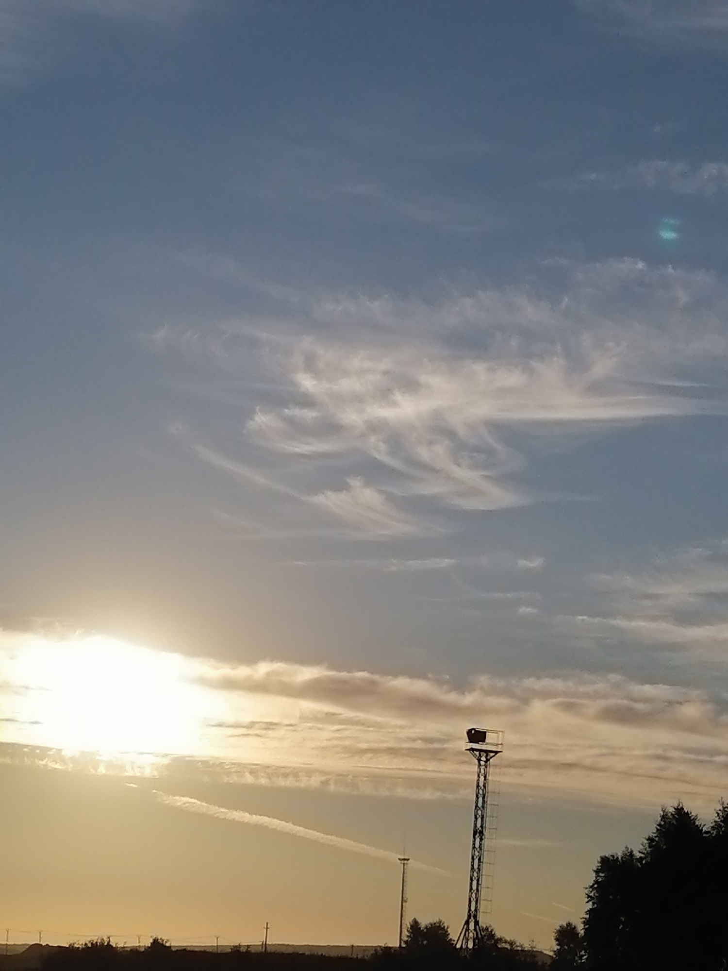 The image depicts a sky with scattered clouds and a bright sun setting on the left side, casting a warm glow. A tall, slender tower with a structure at its peak stands in the lower right quadrant, silhouetted against the sky. There are also trees and a distant horizon line visible. The location is identified as Kharino, Russia.
