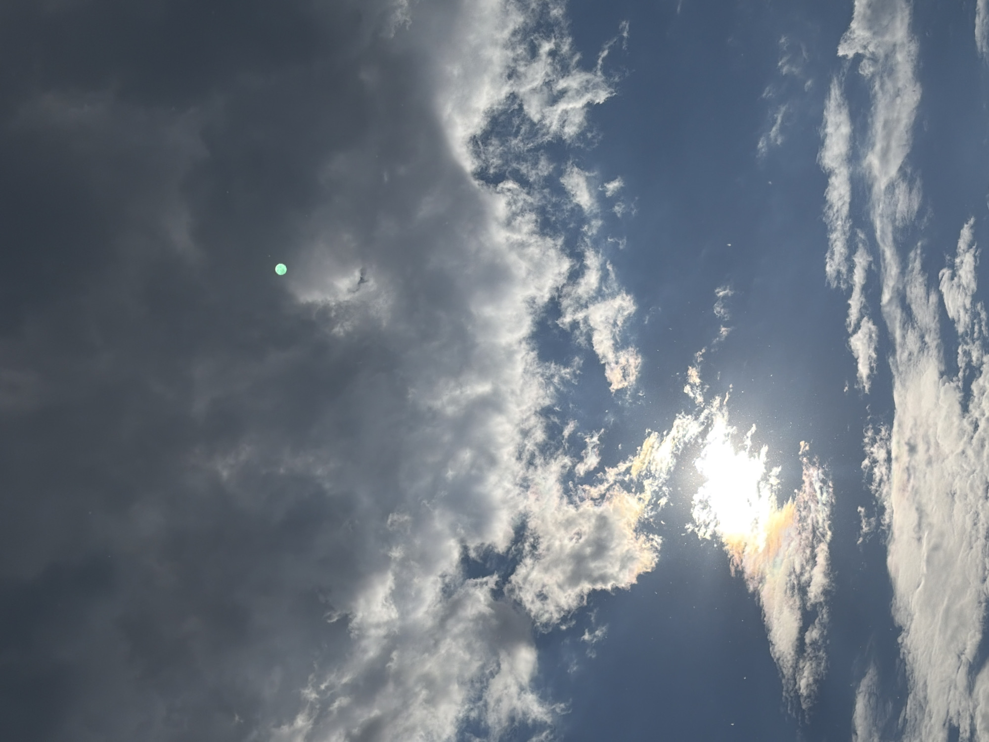 The image captures a sky with dramatic cloud formations and the sun. In the upper portion of the frame, dark, heavy clouds dominate, obscuring the sky. A small, bright green circular object is visible within these darker clouds, appearing to be a lens flare or an anomaly. The lower part of the image shows a bright, intensely lit sun peeking through thinner, wispy clouds. These clouds are illuminated by the sun, creating a colorful iridescence with hues of yellow, orange, pink, and white. Below this, more scattered, white clouds are present against a clear blue sky. There are no people, structures, or specific city landmarks of Chaikovsky, Russia, visible in this frame. The scene is purely atmospheric, focusing on the interplay of light and clouds.