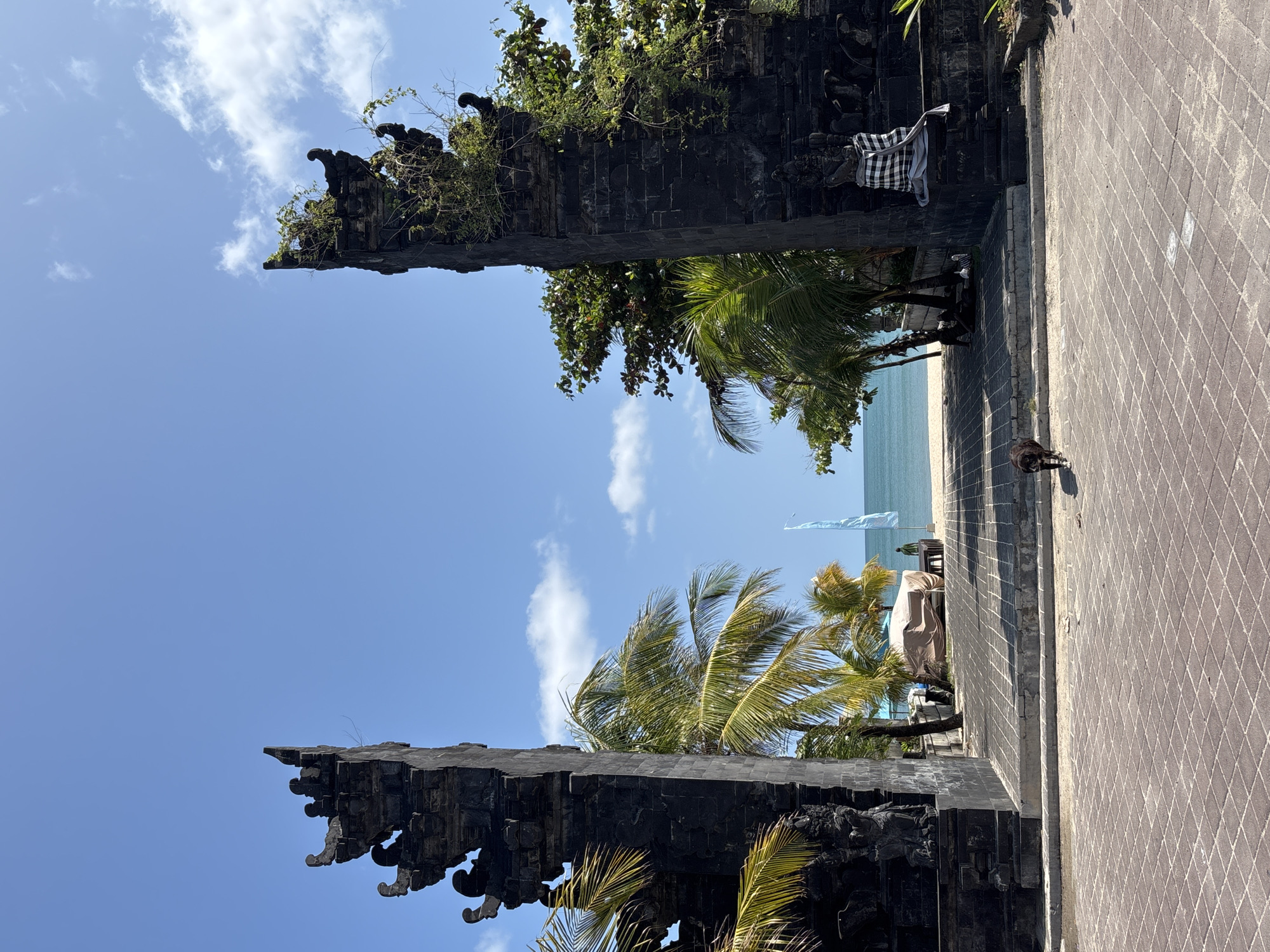 The image shows a view through a traditional Balinese gate in Kuta, Indonesia. The gate is constructed of dark stone, with ornate carvings and vegetation growing on top. Through the gate, palm trees, a beach with light-colored sand, and the ocean are visible. A checkered cloth is draped on one side of the gate. A paved walkway leads through the gate towards the beach. A small dark animal, possibly a dog, stands on the walkway near the right side of the frame. The sky is blue with scattered white clouds. The image is rotated 90 degrees counterclockwise.