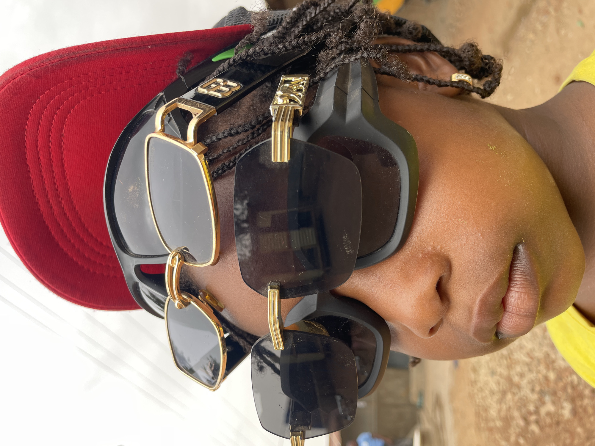 A close-up shot reveals a dark-skinned person with braided hair wearing a red baseball cap and headphones, with two pairs of sunglasses attached to the headphones. The location appears to be outdoors, possibly in the city of Jos, Nigeria. The subject's face is partially visible, focusing on the nose and mouth. A portion of a yellow shirt is visible on the right side of the frame. The background is blurred and indistinct.
