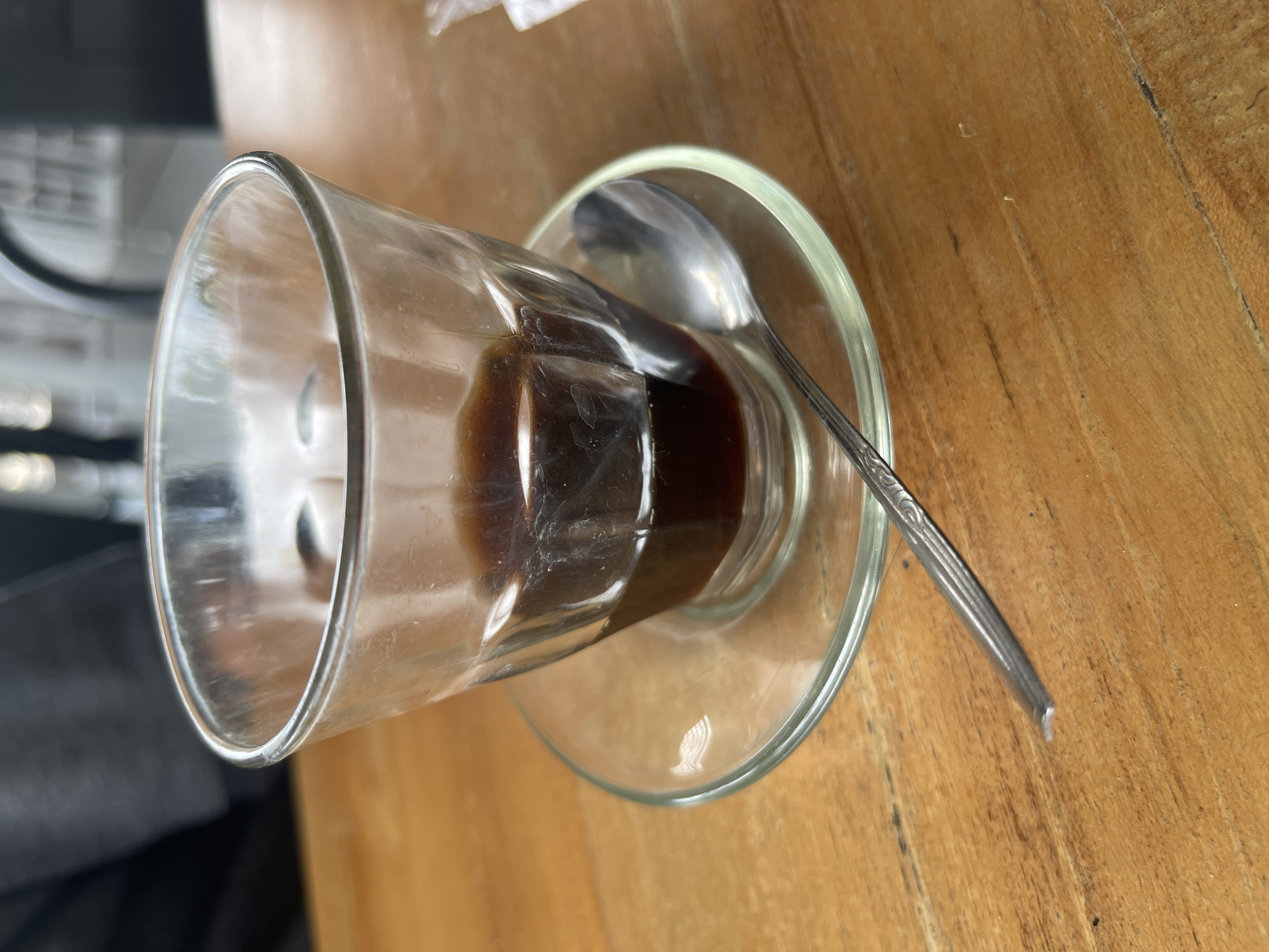 A close-up, eye-level shot shows a clear glass of dark liquid, likely coffee, sitting on a clear glass saucer. A silver spoon rests on the saucer to the right of the glass. The items are placed on a light brown wooden table with visible grain. In the background, slightly out of focus, a person is partially visible wearing dark clothing. The scene appears to be set in an indoor cafe or restaurant in Pulokiton, Indonesia.