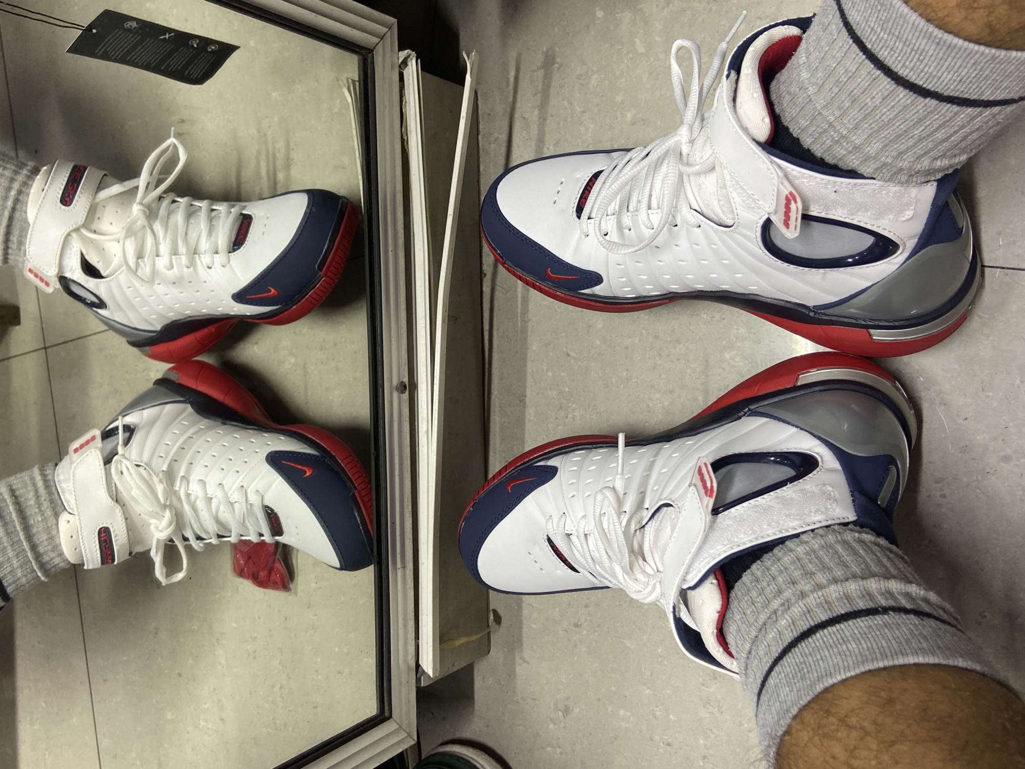 A photograph shows a pair of white, red, and navy blue athletic sneakers being worn by a person with grey socks, reflected in a mirror. The shoes have white laces and velcro straps. The environment appears to be indoors with a concrete floor. The location in the city of Higuerote, Venezuela, is not discernable from the image.