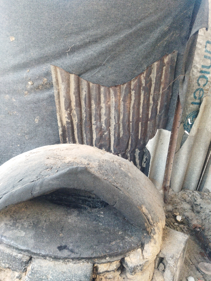 The image displays a weathered, domed outdoor oven, consistent with a tabun, situated in Khan Yunis, Palestinian Territory. The oven's upper portion, constructed from hardened mud or clay, is dark gray, heavily coated with light dust or sand, and shows signs of cracking and erosion around its structure and a dark, empty opening. Its base appears to be built from rough, light-colored bricks or stones, also covered in dust.
Behind the oven, an improvised backdrop or wall is visible. This includes a large sheet of dark gray fabric, likely canvas or tarp, which is creased, dusty, and appears draped or stretched. Partially visible through or behind this fabric is a section of rusted, corrugated metal sheeting, characterized by vertical ridges and streaks of lighter material. To the right of the corrugated metal, a lighter-colored sheet of material is present, displaying the word "UNICEF" in blue, vertically oriented text. Further to the right, additional pieces of irregularly shaped, light-colored debris are visible.
The ground immediately surrounding the right side of the oven is unpaved earth or sand, containing small stones and accumulated debris. A vertical, dark metal rod extends upwards from the ground near the oven's right side. No individuals, explicit actions, or interactions are present in the frame.