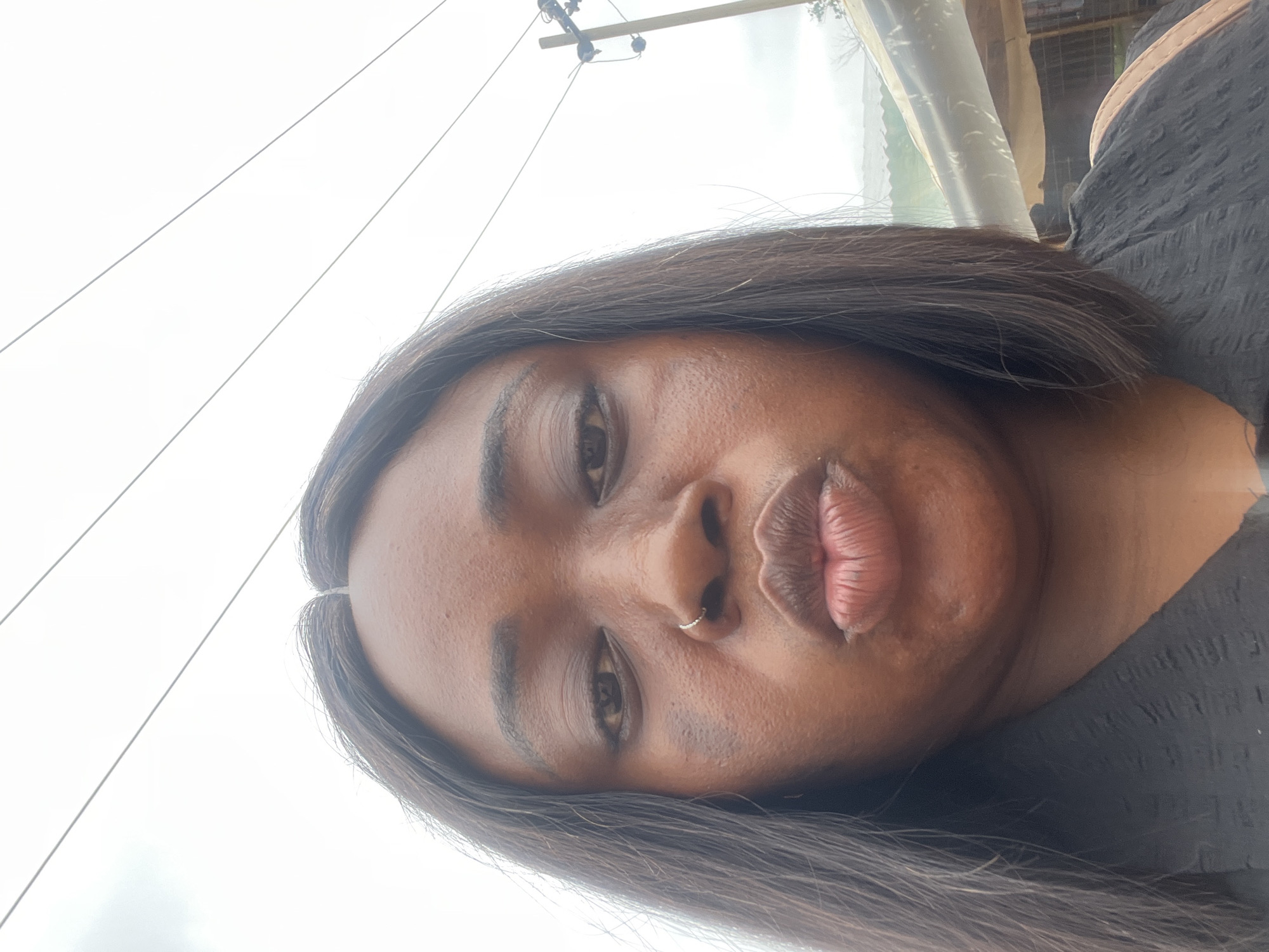 A dark-skinned woman with short, dark brown hair is facing the camera. She has a nose ring in her left nostril. The background includes power lines, a utility pole, and what appears to be a partially covered structure. The location is presumed to be Abuja, Nigeria.