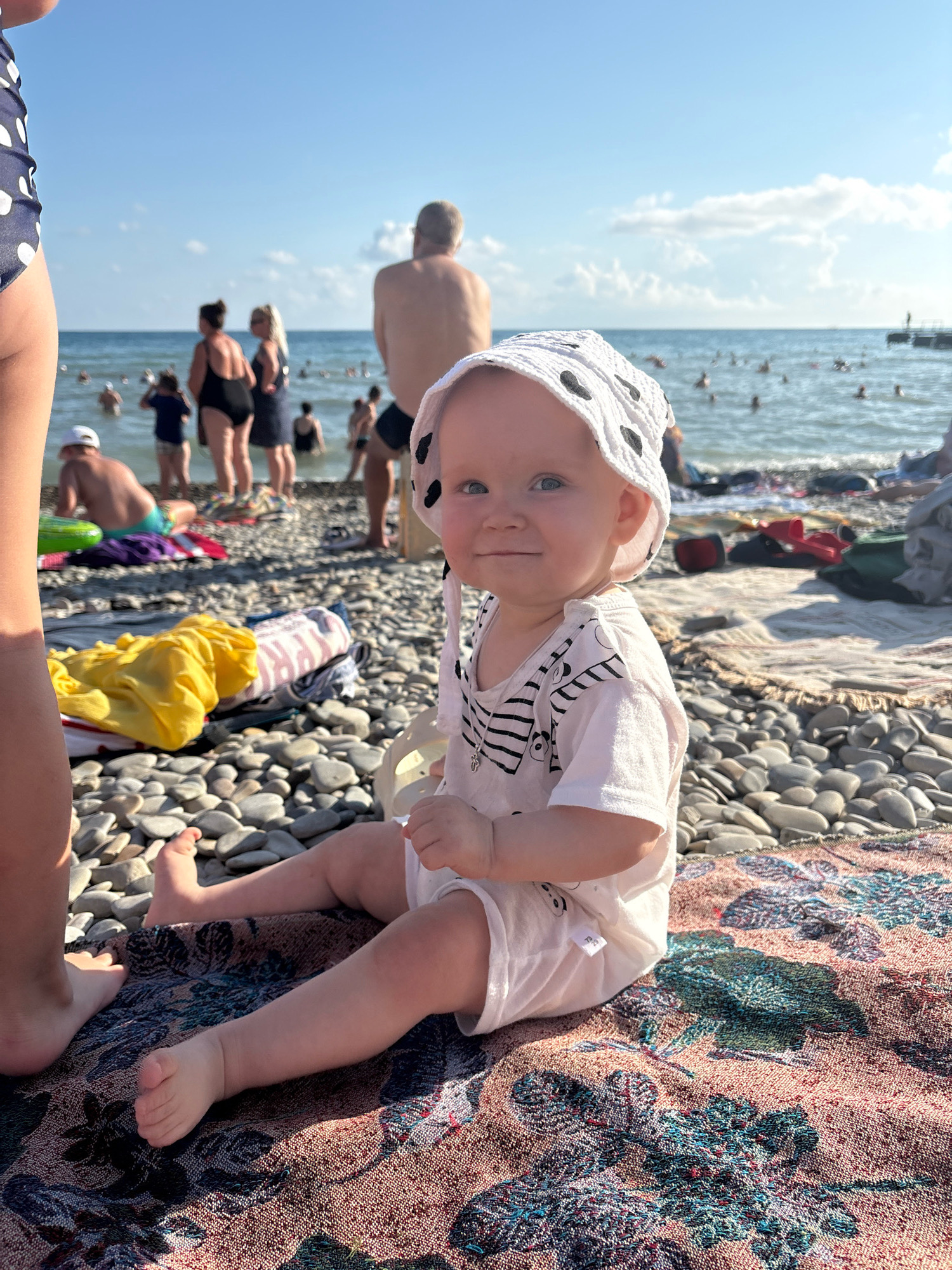 The image depicts a bright, sunny day at the beach in Arkhipo-Osipovka, Russia. In the foreground, a baby wearing a white outfit and a patterned hat sits on a colorful blanket spread across a pebble beach. The baby is looking towards the camera with a slight smile. To the left, the legs of another person are visible, partially cut off by the frame. The beach is populated with various individuals, some in the water and others sunbathing or sitting on towels. The sea is calm, and the sky is a clear blue with a few scattered clouds. In the background, a pier extends into the water, and people can be seen enjoying the beach and the sea. Several beach towels and bags are scattered around, indicating a leisurely day at the seaside. The overall scene conveys a relaxed and cheerful atmosphere.