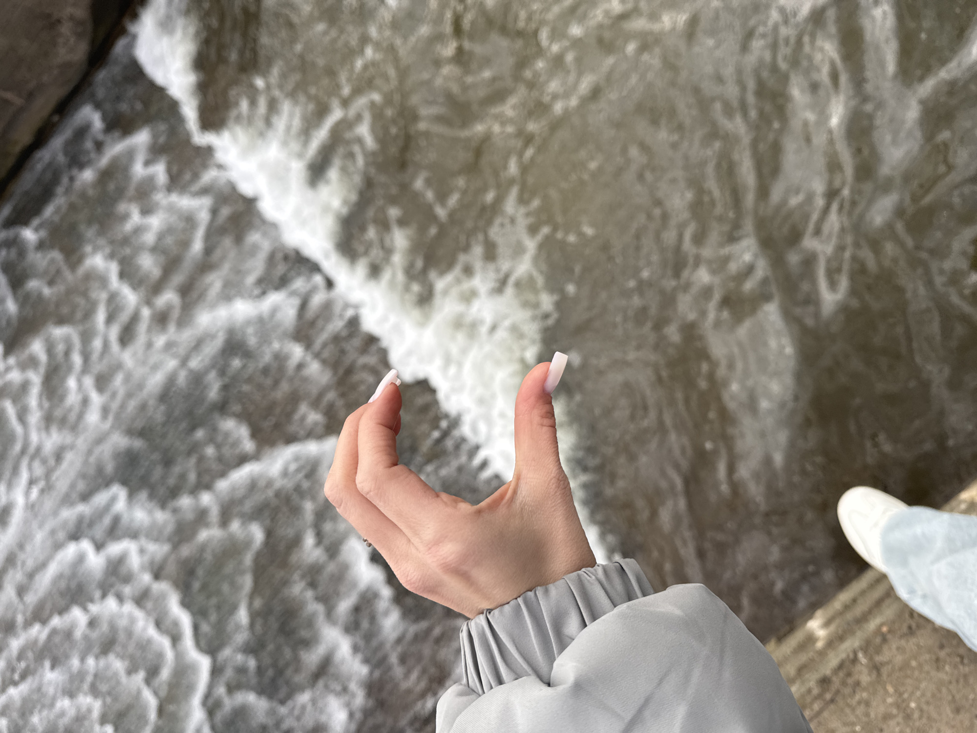 An overhead shot captures a person's right hand and forearm extending over a turbulent body of water. The hand, with light skin tone and long, light-colored square-tipped nails, is positioned in a semi-cupped or 'c'-shape, with the thumb and index finger raised. The person wears a grey jacket with a gathered, elasticated cuff. The water below is a mix of dark grey and foamy white, indicating rapid movement, possibly a river or stream with strong currents or rapids. The water in the foreground appears more churned and white, while further back it is darker and more fluid, with visible swirls. In the bottom right corner, a portion of a light-colored shoe and light blue pants or jeans are visible, belonging to the same person. The overall lighting is soft and diffused, suggesting daytime without direct sunlight. There is no visible text in the image. The scene is set in what appears to be a natural outdoor environment, possibly in or near Фоки, Russia.