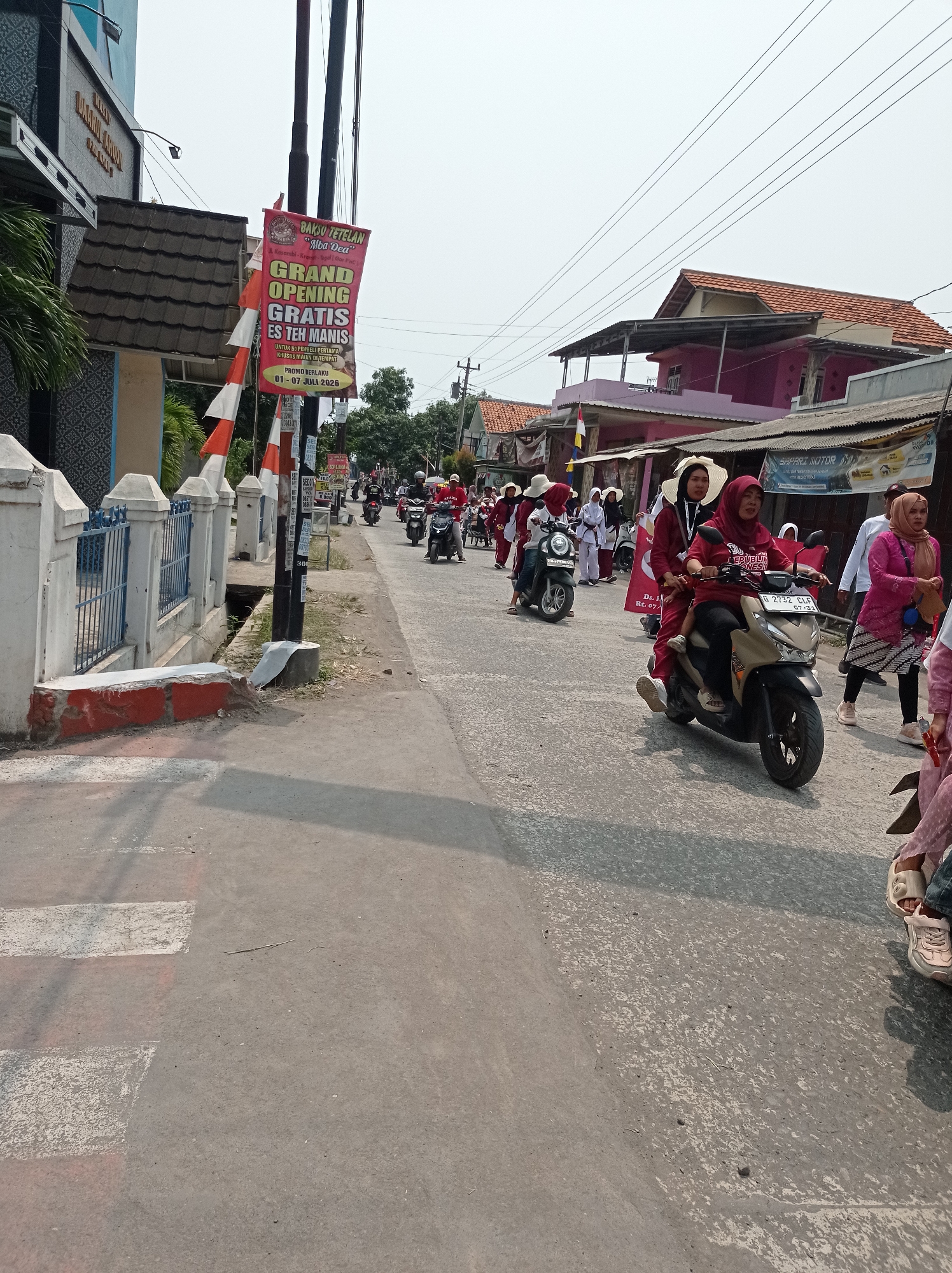 The image depicts a street scene in Kramat, Indonesia. A banner for "Bakso Tetelan 'Alba Dea'" advertises a grand opening with free sweet iced tea for the first 50 customers, valid from July 1st to July 7th, 2026. A procession of people on motorcycles, many wearing red and white attire and some with hijabs, moves along the street. A woman on the front motorcycle wears a red shirt with "REPUBLIK INDONESIA" printed on it. The road is paved, and there is a pedestrian crossing marked by white stripes. Buildings line the street, with one featuring a blue and white patterned facade and another with a pink exterior. Power lines are visible overhead. The overall atmosphere suggests a festive or celebratory event.