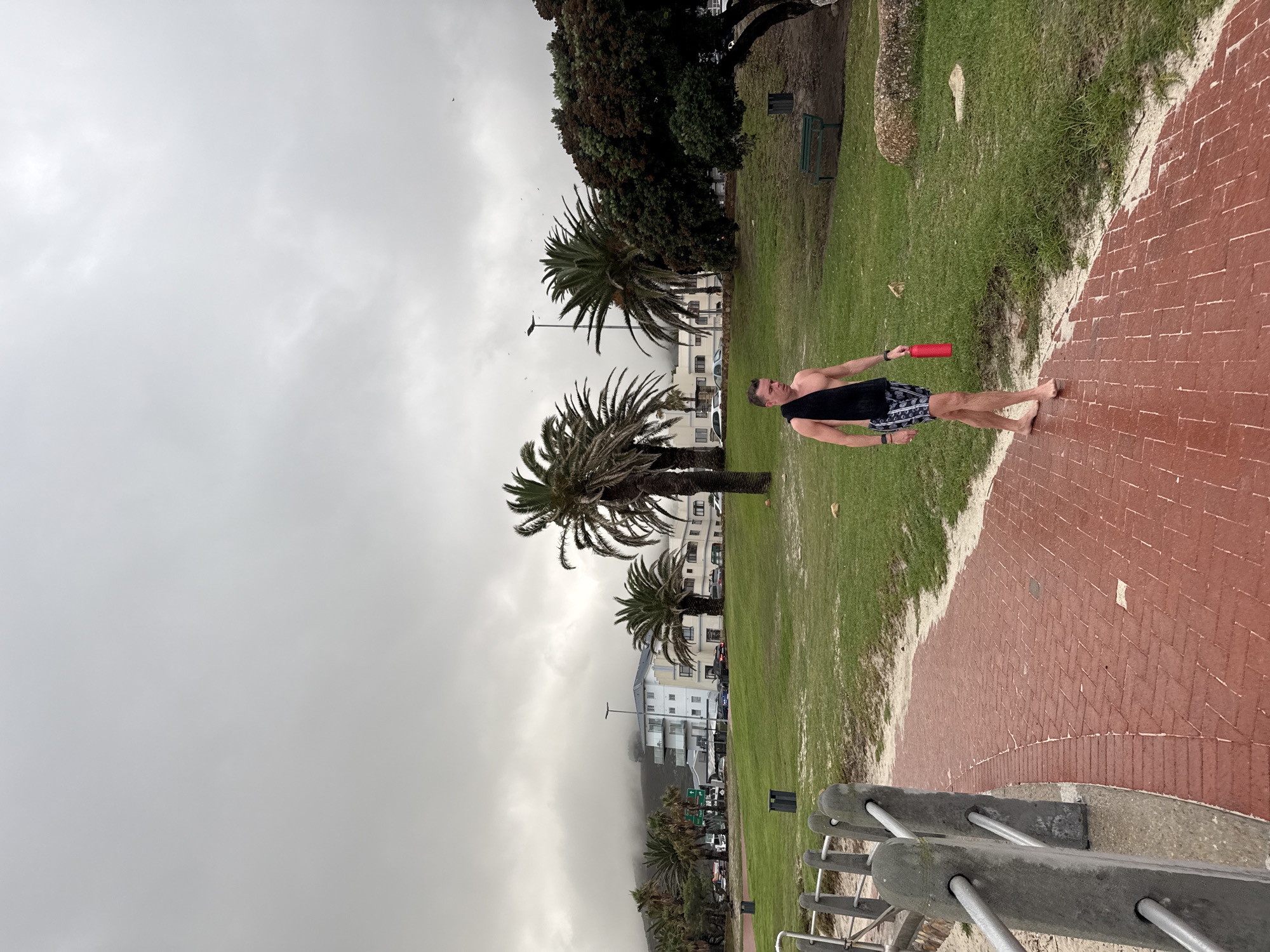 A medium shot, taken outside in daylight in Cape Town, South Africa, shows a Caucasian male walking barefoot across a brick pavement area towards a grassy area with palm trees. The man is wearing patterned shorts and a black, sleeveless top. He is holding a red object in his right hand. The backdrop features buildings shrouded in fog, suggesting a cloudy or overcast day. A metal railing is visible in the foreground. The overall setting appears to be a park or public space adjacent to the coastline, typical of Cape Town's coastal environment.