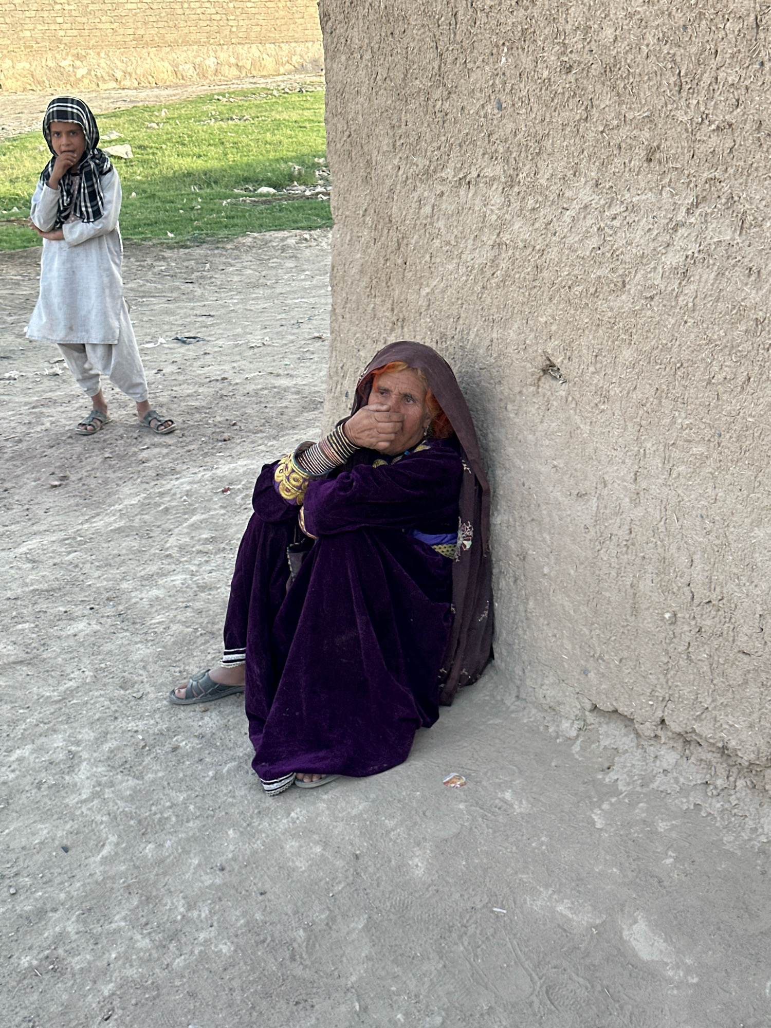 In Kuchlak, Pakistan, an elderly woman sits slumped against a rough, textured, light brown wall, her body angled towards the right side of the frame. She is wearing a dark purple velvet garment and a dark brown headscarf that covers her hair and drapes around her face, obscuring her left side. Her right hand is raised, covering her mouth and nose. She is adorned with numerous colorful bangles and a gold bracelet on her right wrist. Her left foot, clad in a gray sandal, rests on the ground, while her right foot is not visible.
To the left and further back, a young person, likely a child, stands facing towards the viewer. They are wearing a light-colored tunic and pants, and a black and white checkered scarf is wrapped around their head and neck. Their left hand is raised to their chin, and their right arm is bent at the elbow, held across their chest. This child also wears gray sandals.
The ground beneath them is a coarse, light gray dirt or gravel, with some scattered debris. Behind the child, a patch of green grass is visible, bordered by a low, light brown brick wall. The overall environment suggests an outdoor, dusty setting, possibly in a rural or semi-urban area.