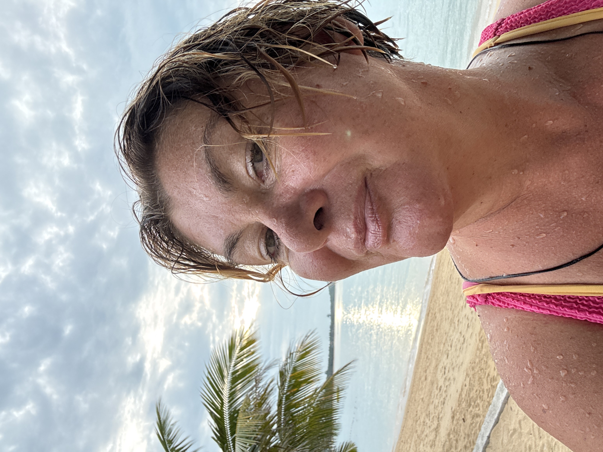 A close-up, slightly angled portrait of a woman standing on a beach in Baan Bo Phut, Thailand. The woman has short, wet hair and is covered in water droplets. She wears a swimsuit with pink and yellow straps. The background includes a palm tree, the ocean, a sandy beach, and a cloudy sky. The sun is reflecting off the water.