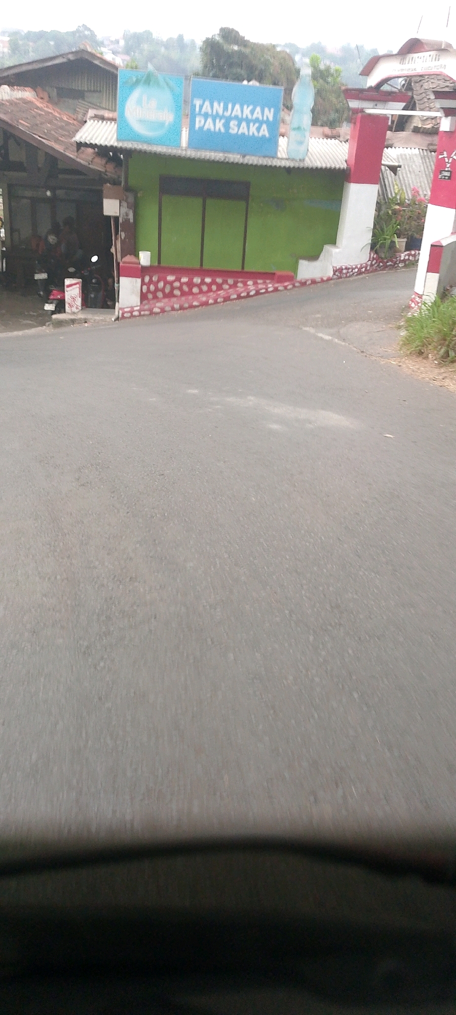 This is a medium-angle, eye-level outdoor shot of a road leading towards a gateway structure in Bandung City, Indonesia. The road is paved and slopes slightly upwards. On the right side of the road, there is tall, dry grass.
The gateway structure is prominent, featuring red and white vertical pillars. It appears to be an entrance to a community or a specific area. Behind the gateway, a green building with a white corrugated metal roof is visible. Above the green building, two signs are mounted on the roof. The left sign is blue with a circular graphic and text, and the right sign is a rectangular blue sign with white text that reads "TANJAKAN PAK SAKA". A large, clear plastic bottle of water is placed on top of the roof between the two signs.
To the left of the green building, there is a structure with a weathered, dark-colored roof and a section of white corrugated metal roofing. Underneath this structure, several motorcycles are parked, and two people are visible in the shadows. A red and white striped bollard is positioned near the motorcycles.
The foreground of the image is dominated by the road, with the bottom portion of the frame blurred, suggesting the photo was taken from a moving vehicle. The overall impression is that of a rural or semi-urban setting in Bandung, Indonesia, possibly marking an entrance to a neighborhood or a local business.