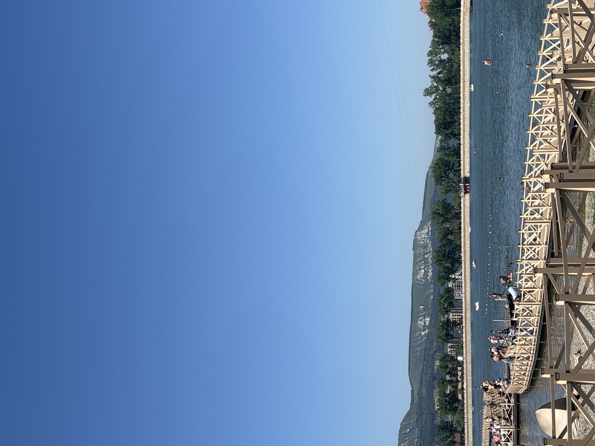 The image depicts a sunny day at a recreational lake in Kislovodsk, Russia. In the foreground, a wooden pier with light-colored railings curves into the water, with several people standing and walking on it. Some are dressed in swimwear. The lake is populated with swimmers and a few small boats. Across the water, a tree-lined embankment separates the lake from a town or resort area, characterized by multi-story buildings and a distant mountain range. The sky is a clear, bright blue.