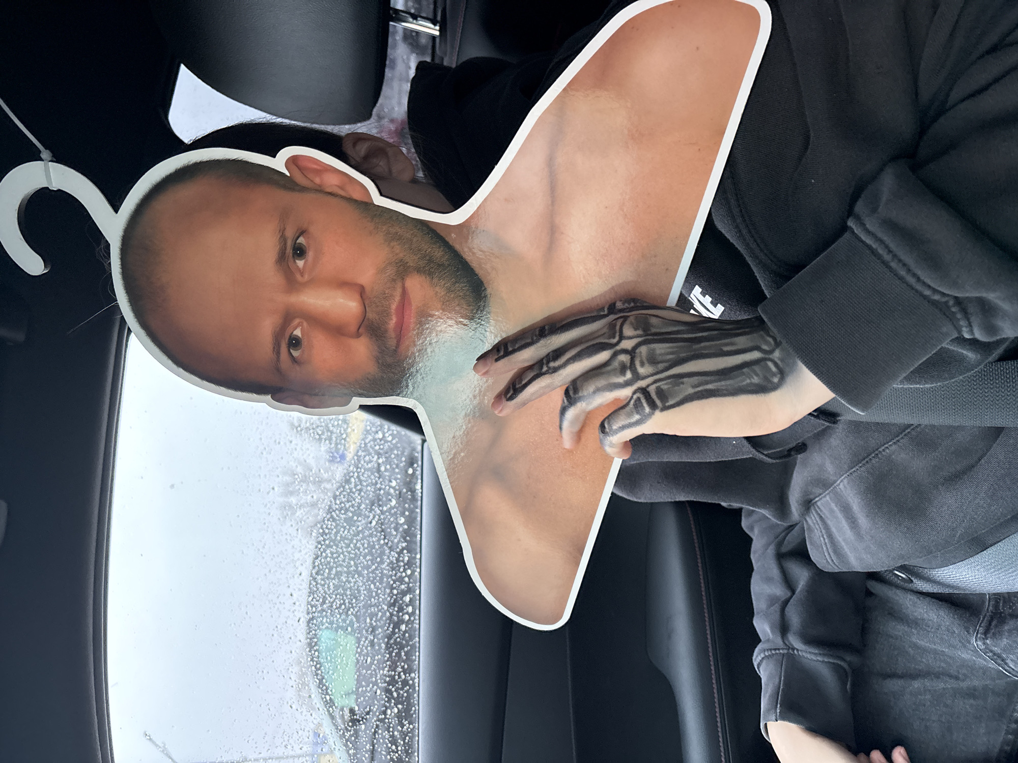 A person sits in the passenger seat of a car. The car is likely parked, judging by the lack of motion blur and the visible raindrops on the window. The person holds up a cutout depicting a man's head and upper torso with a skeletal hand superimposed. The cutout is attached to a white plastic clothes hanger. The person's clothing consists of a dark-colored hoodie or sweatshirt. The location is inferred to be Chaikovsky, Russia, from the prompt's instructions. No specific features in the image definitively confirm the city, but the assumption is made based on the provided context.