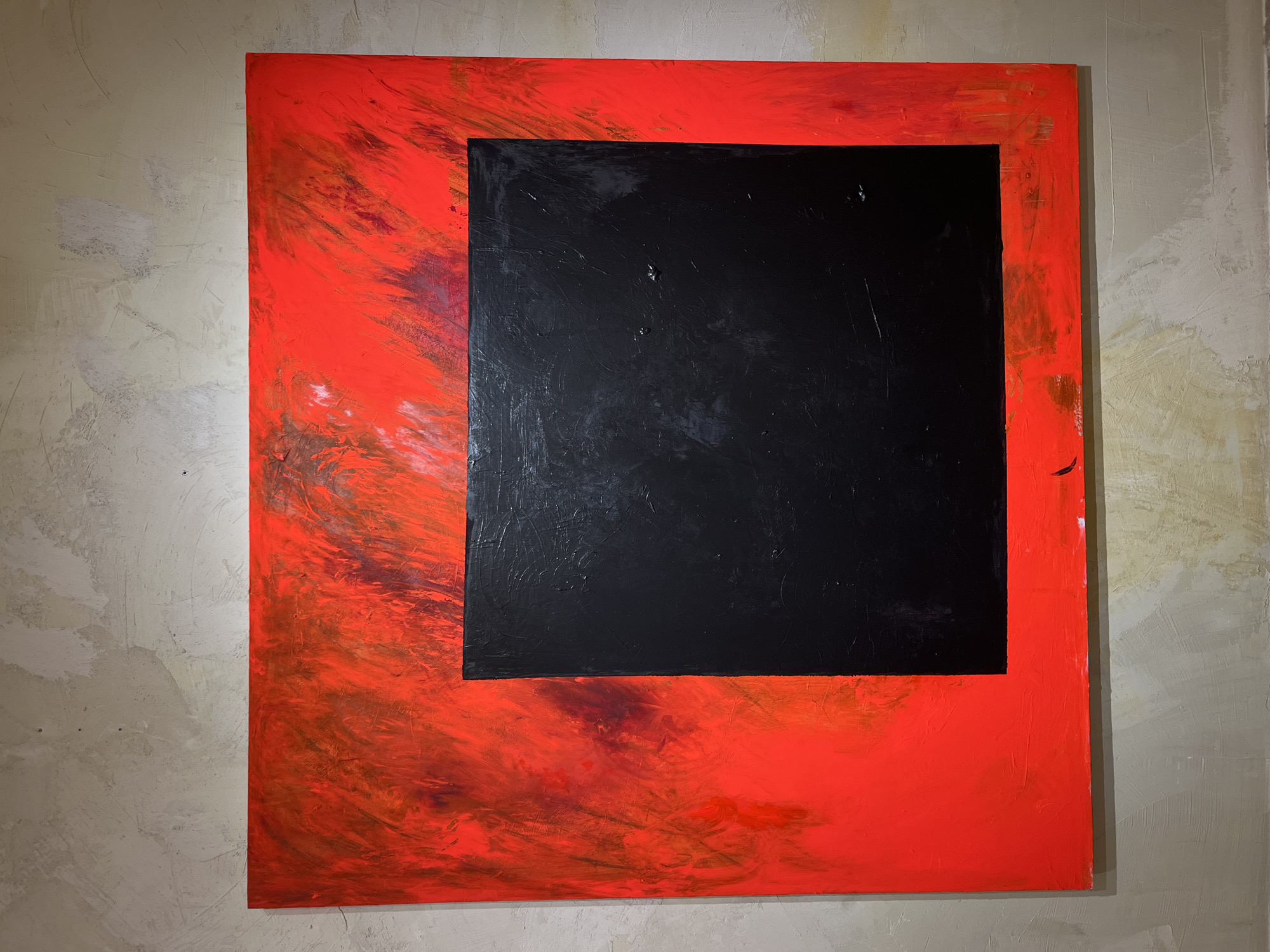 The image shows a painting with a black rectangle in the center, surrounded by a vibrant red border. The red area is textured and appears to have brushstrokes of various shades of red and some darker tones. The painting is hung on a textured, light-colored wall. The wall has an uneven, stucco-like finish. The lighting suggests an indoor setting, possibly a gallery or an exhibition space. There are no people or animals visible in the image. The painting itself is the sole focus. The exact location in Barcelona, Spain, cannot be determined from the image alone, as it depicts an artwork and its immediate surroundings, without any identifying landmarks or contextual clues specific to Barcelona.