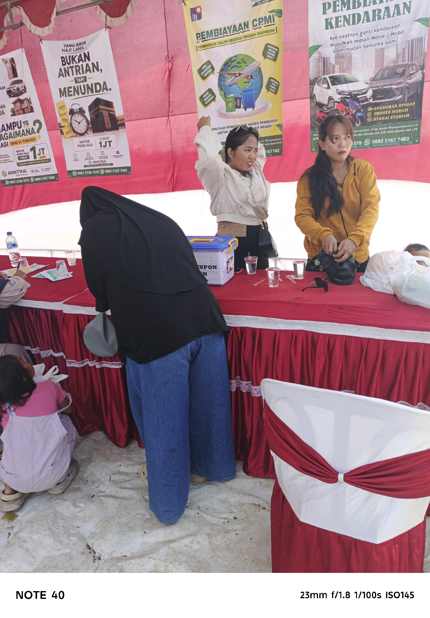 In Desa Lomanis, Indonesia, an outdoor scene unfolds with several people gathered around a table draped in a red tablecloth. The table is set up for an event, with promotional banners hanging in the background. One banner advertises "PEMBIAYAAN DARI KENDARAAN" and features images of cars and motorcycles. Another banner promotes "PEMBIAYAAN KPKM" with graphics of a globe and luggage, suggesting travel or migrant worker services.
A young woman in a yellow hoodie stands behind the table, interacting with a black handbag. To her left, another woman in a white jacket with her arm raised appears to be speaking. In front of the table, a person wearing a black hijab and loose blue jeans is bent over, facing the table. To the left, a young child in a pink outfit is squatting on the ground. Several plastic cups and what appear to be promotional flyers are placed on the table. The ground is covered with a white tarp, suggesting an improvised event space. The overall atmosphere suggests a public event or service booth.
