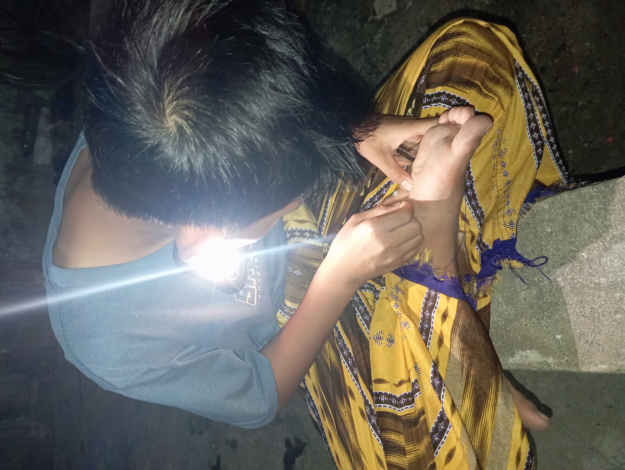 A person is sitting on a concrete ledge in Rejosari, Indonesia, illuminated by a bright light source. They are wearing a light blue t-shirt and a yellow and brown patterned sarong. The person is bent over, examining their left foot with their hands. Their hair is dark and appears somewhat disheveled. The environment is dimly lit, suggesting it might be nighttime or indoors with artificial lighting.