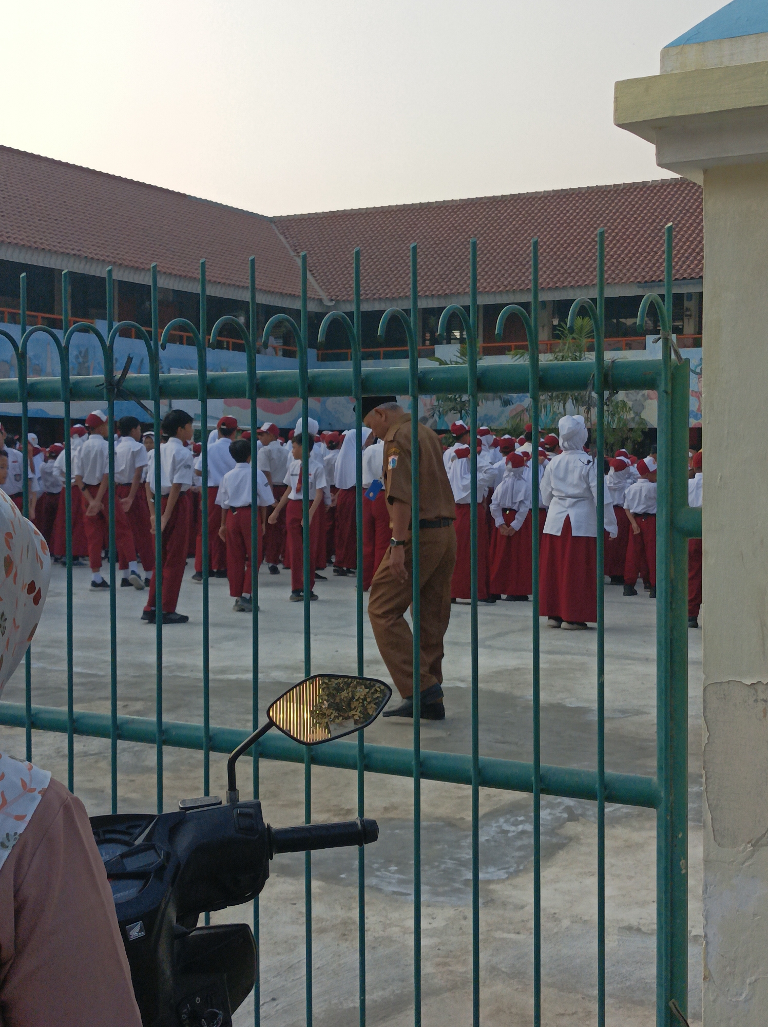A group of schoolchildren in white shirts and red skirts or pants stand in an outdoor courtyard. The children appear to be in formation, possibly for a school assembly or activity. A male adult, wearing a brown uniform and a hat, stands facing away from the camera, with his hands clasped behind his back. He is looking towards the children. The scene is viewed through a green metal fence, which partially obscures the view. In the foreground, a motorcycle is partially visible on the left, and a portion of a woman's headscarf is on the far left. The background shows a building with a red tiled roof and a blue painted wall with some murals. The sky is pale and overcast. The location is identified as a school in West Jakarta, Indonesia.