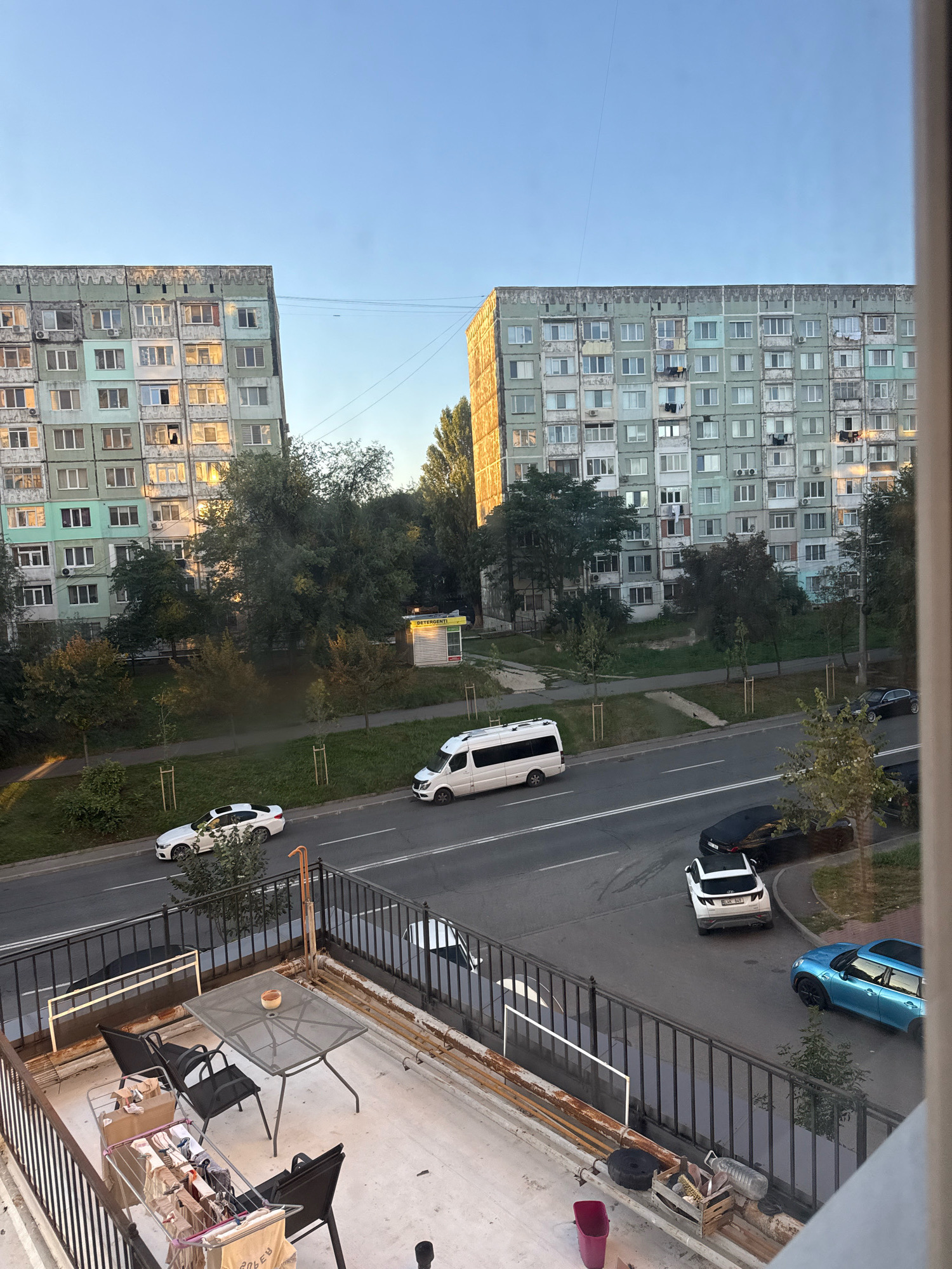 The image depicts a street scene in Chișinău, Moldova, captured from a balcony. In the foreground, a concrete balcony is furnished with a metal table and four chairs. A clothes rack with laundry is also present. The balcony overlooks a street with several cars, including a white minivan, a white sedan, a white SUV, a black sedan, and a blue hatchback. Two large, aging apartment buildings, characteristic of Soviet-era architecture, dominate the background. Trees and a small kiosk with the word "DETERGENTI" are visible between the buildings and the street. The sky is clear and blue, indicating daytime. The overall scene suggests a residential area in Chișinău.