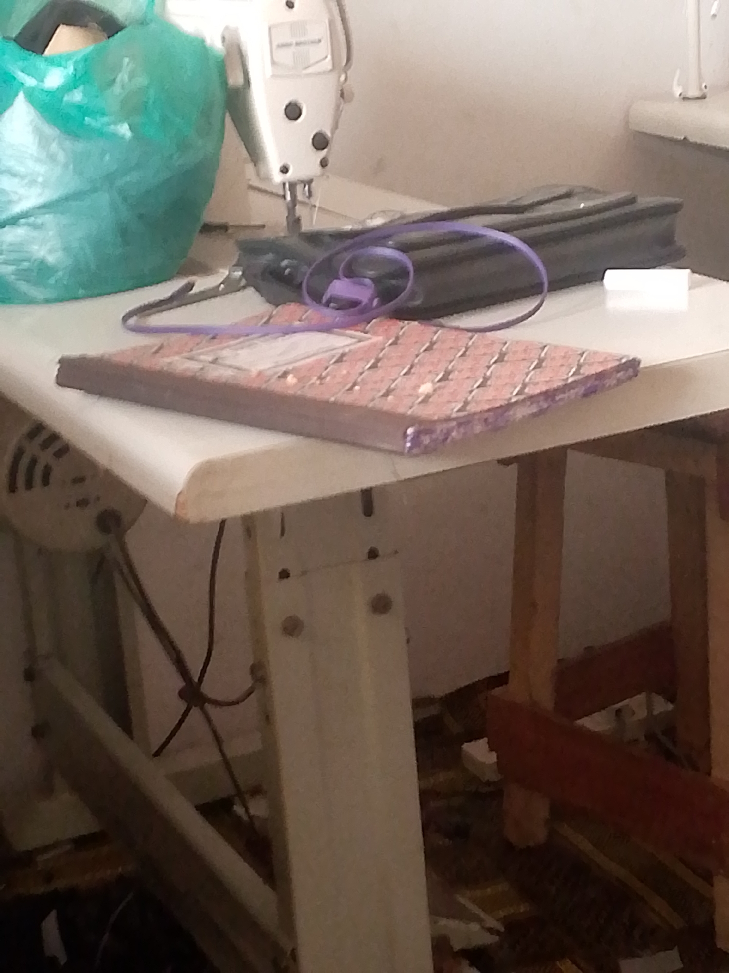 The image shows a close-up of a white sewing machine on a table. To the left of the sewing machine is a crumpled teal plastic bag. A black zippered bag is lying on the table behind a pink and purple patterned notebook. A white rectangular object is placed in front of the black bag. The sewing machine has a needle and presser foot lowered. A purple string is looped around the notebook and the presser foot of the sewing machine. The table appears to be made of white laminate, and the sewing machine is mounted on a metal stand. The background is out of focus and shows a wall and a wooden chair leg. The scene is indoors, possibly a workshop or a sewing room. There are no people visible in the image. The photograph does not contain enough information to determine the specific location within Jalingo, Nigeria.