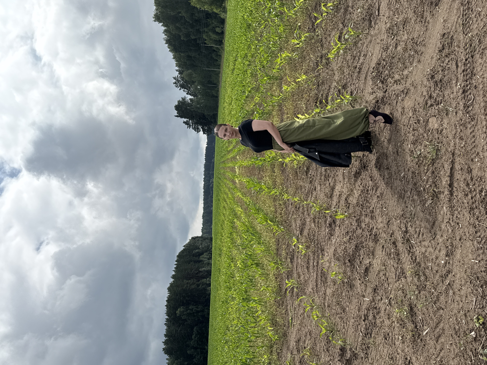 A woman stands in a cornfield in the city of Prikamsky, Russia, under a cloudy sky. She is wearing a black t-shirt, a long olive-green skirt, and black heels. She is holding a black jacket. The cornfield is in the foreground and extends into the background, bordered by a line of trees. The sky is overcast with dramatic clouds. The ground is dirt with visible tire tracks.