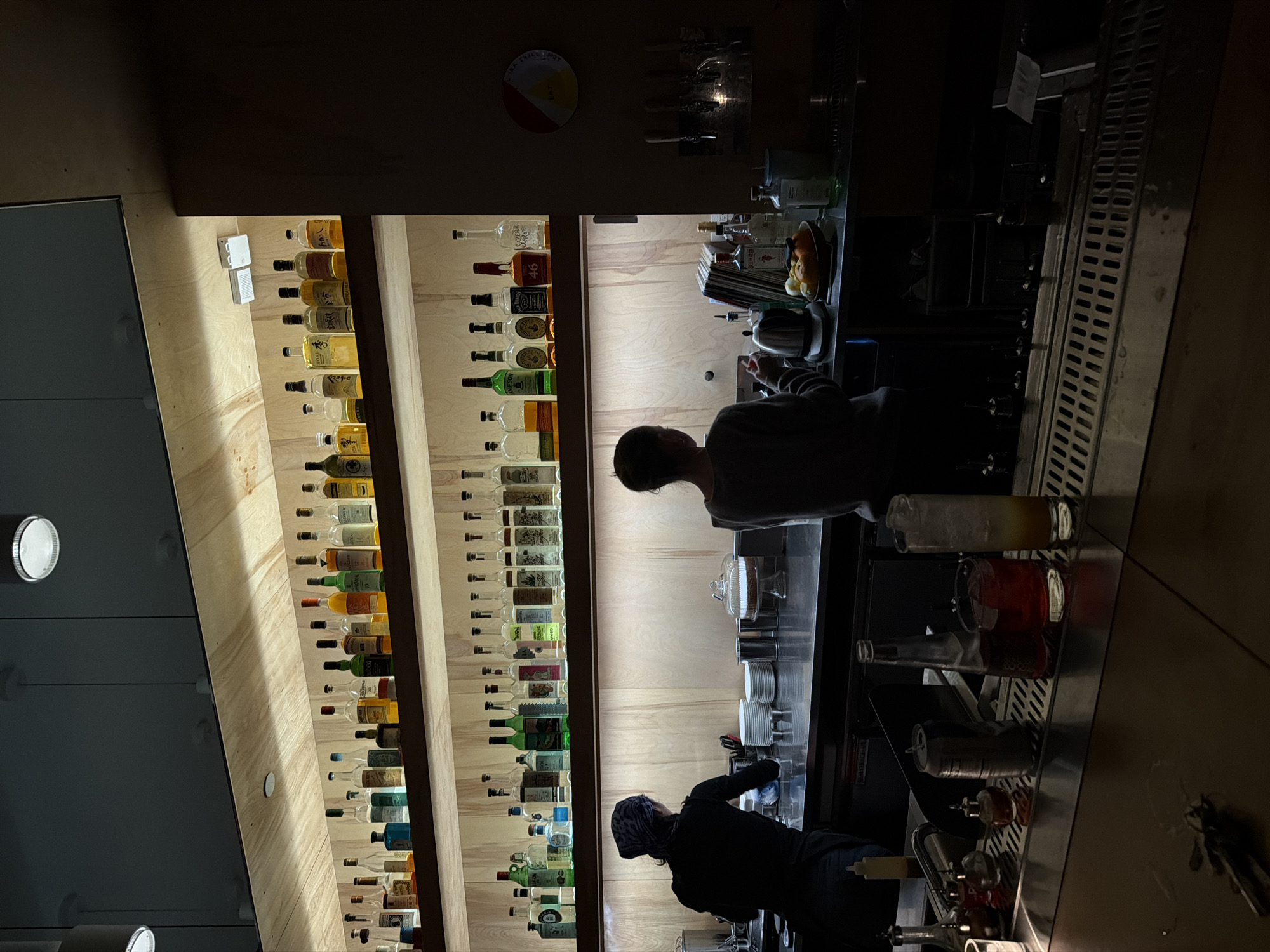 This image shows the interior of a bar. Two people are visible; one is wearing a light grey shirt and the other is wearing a dark garment with a bandana. They appear to be working behind the bar. The shelves behind them are filled with various bottles of alcohol. On the bar top, there are glasses, bottles, and what looks like bartending tools. The environment appears dimly lit. I am unable to identify the specific location within New York City.