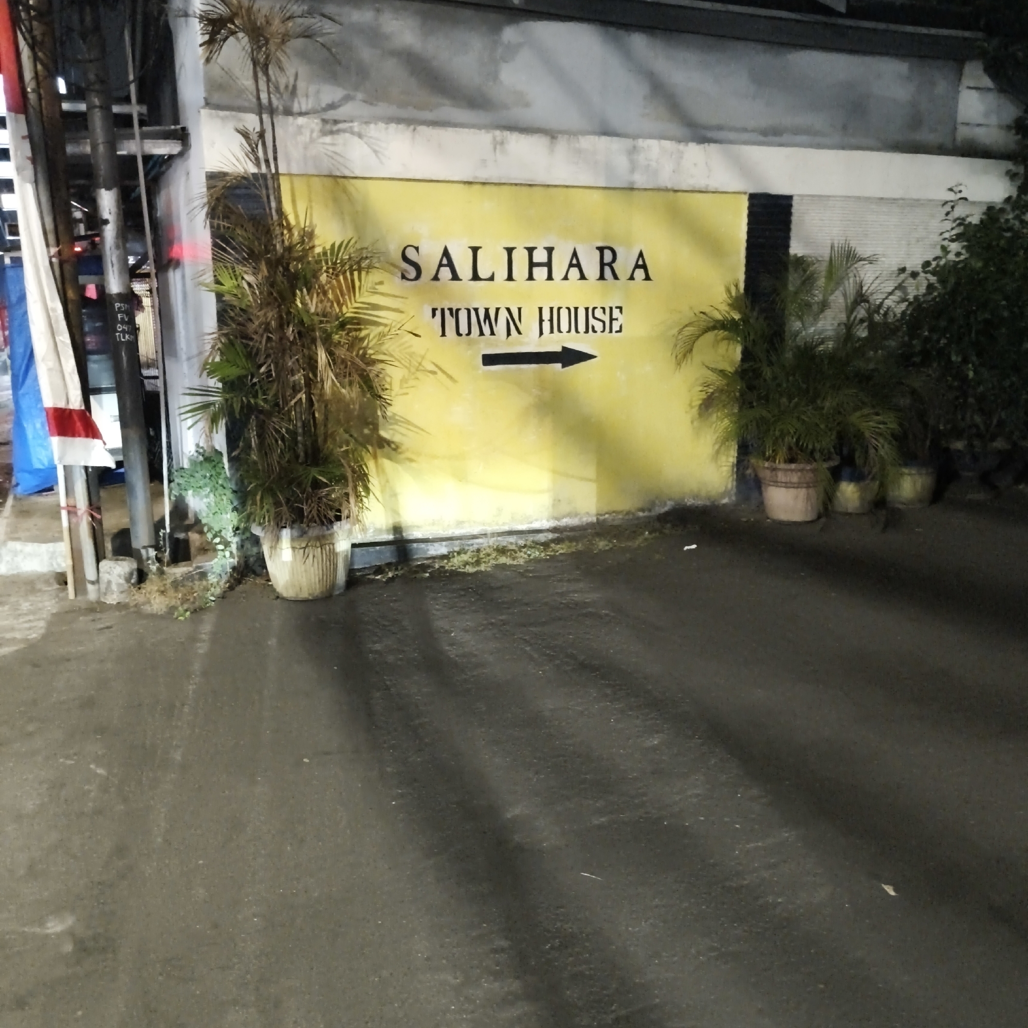 This is a medium shot, eye-level, outdoor photograph of the entrance to "SALIHARA TOWN HOUSE" in South Jakarta, Indonesia. The building facade is painted a bright yellow, with the name of the establishment written in black sans-serif font above an arrow pointing to the right. To the left of the entrance are several utility poles with wiring, a white and red flag, and a blue tarp. In front of the building, to the left and right, are large potted plants. The ground is wet and appears to be asphalt, suggesting it may have recently rained or the area is being watered. Shadows are cast on the ground from the plants and poles, indicating the lighting conditions. The overall scene depicts an entrance to a residential or commercial property.