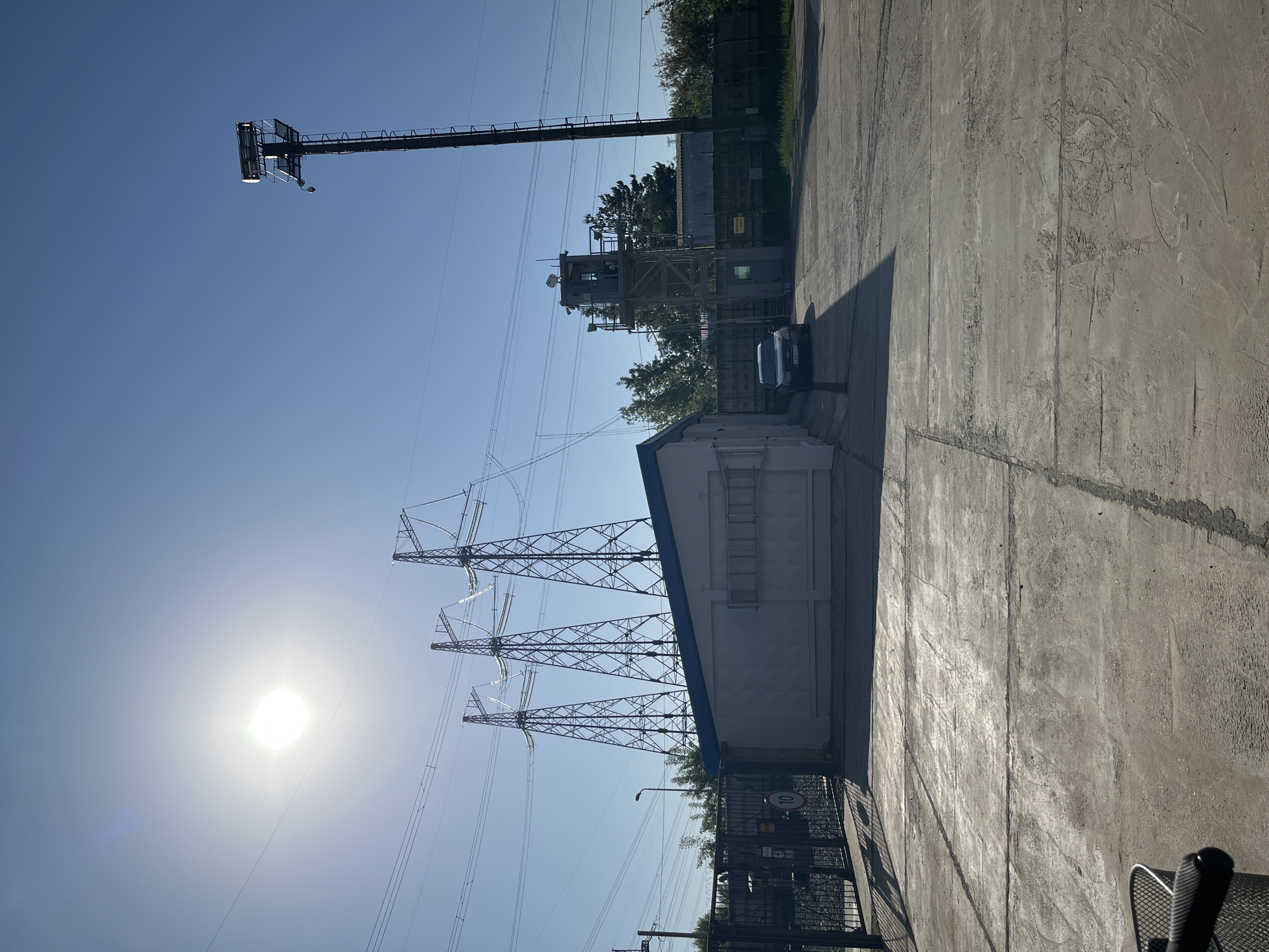 The image depicts an industrial area in the city of Chaikovsky, Russia, characterized by power infrastructure and utilitarian architecture. In the foreground, a concrete surface extends towards the right, showing signs of wear. A tall metal fence and gate, with a posted speed limit sign, separate this area from a white rectangular building with a blue roof, featuring a metal ladder on its side.
Three steel lattice-type high-voltage power line towers rise into the blue sky, supporting electrical transmission lines. In the background, behind the building, a dark-colored car is parked near what appears to be a metal observation or control tower. Further in the distance, a tall metal mast, possibly a communication or lighting tower, is visible. The sun is positioned in the lower left, casting bright light and shadows across the scene. There are no people visible. The environment is industrial with a clear sky.