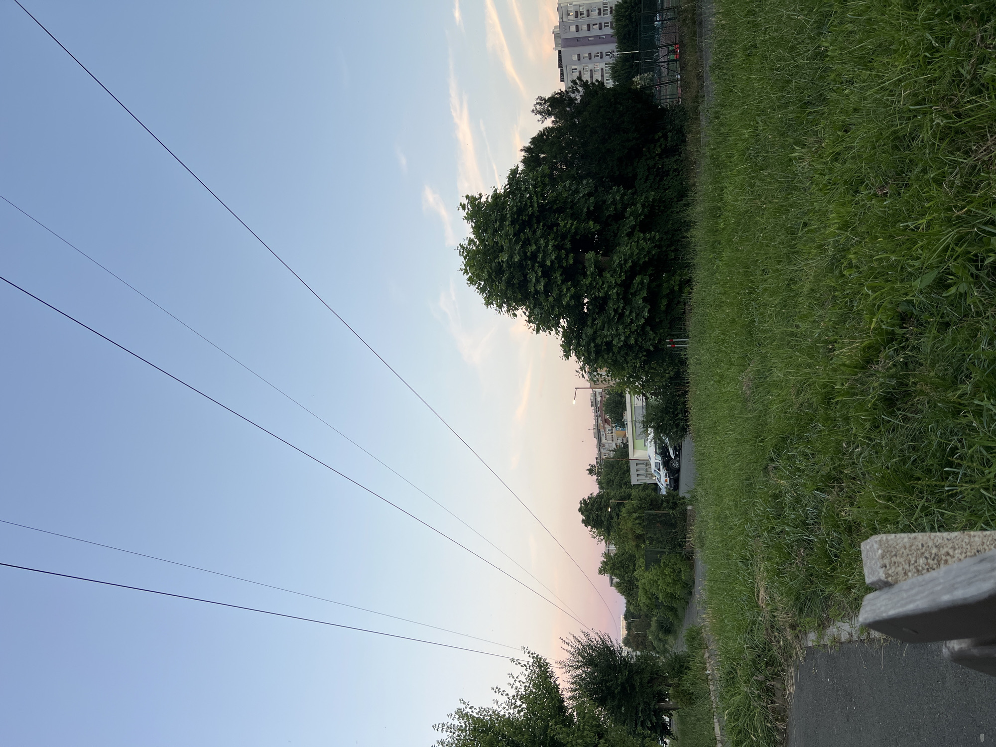 The image shows a suburban landscape in Burgas, Bulgaria, likely during dusk. Four power lines stretch diagonally across the pale blue sky, which is tinged with pink near the horizon. In the foreground, there is a patch of tall, uncut grass and a paved pathway. A portion of a grey wooden bench with concrete supports is visible in the lower left corner. Beyond the grass, a line of trees and bushes separates the area from a parking lot where several white cars and a dark SUV are parked. In the background, apartment buildings with multiple windows are visible, suggesting a residential area. To the right, a fenced sports court can be seen. The overall impression is one of a quiet, everyday scene in a city.