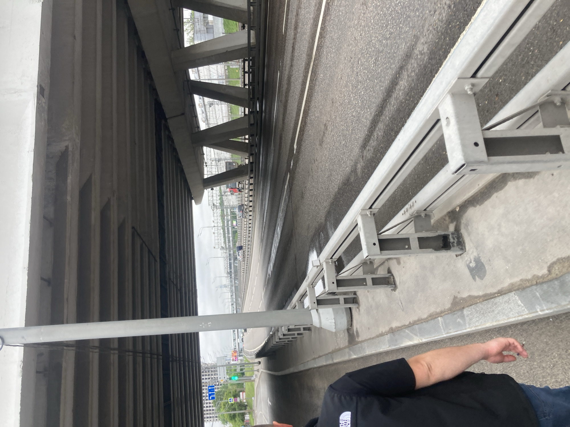 The image shows a view from under a bridge structure, alongside a road in Khimki, Russia. The bridge is made of concrete and steel with multiple support beams. The road is asphalt with painted lane markings and a metal guardrail on the right side of the frame. Traffic is visible on the road in the distance. A person, partially visible in the lower right corner, is wearing a black shirt and blue jeans. Green vegetation is seen on the far side of the road. Signage and buildings are visible in the background.