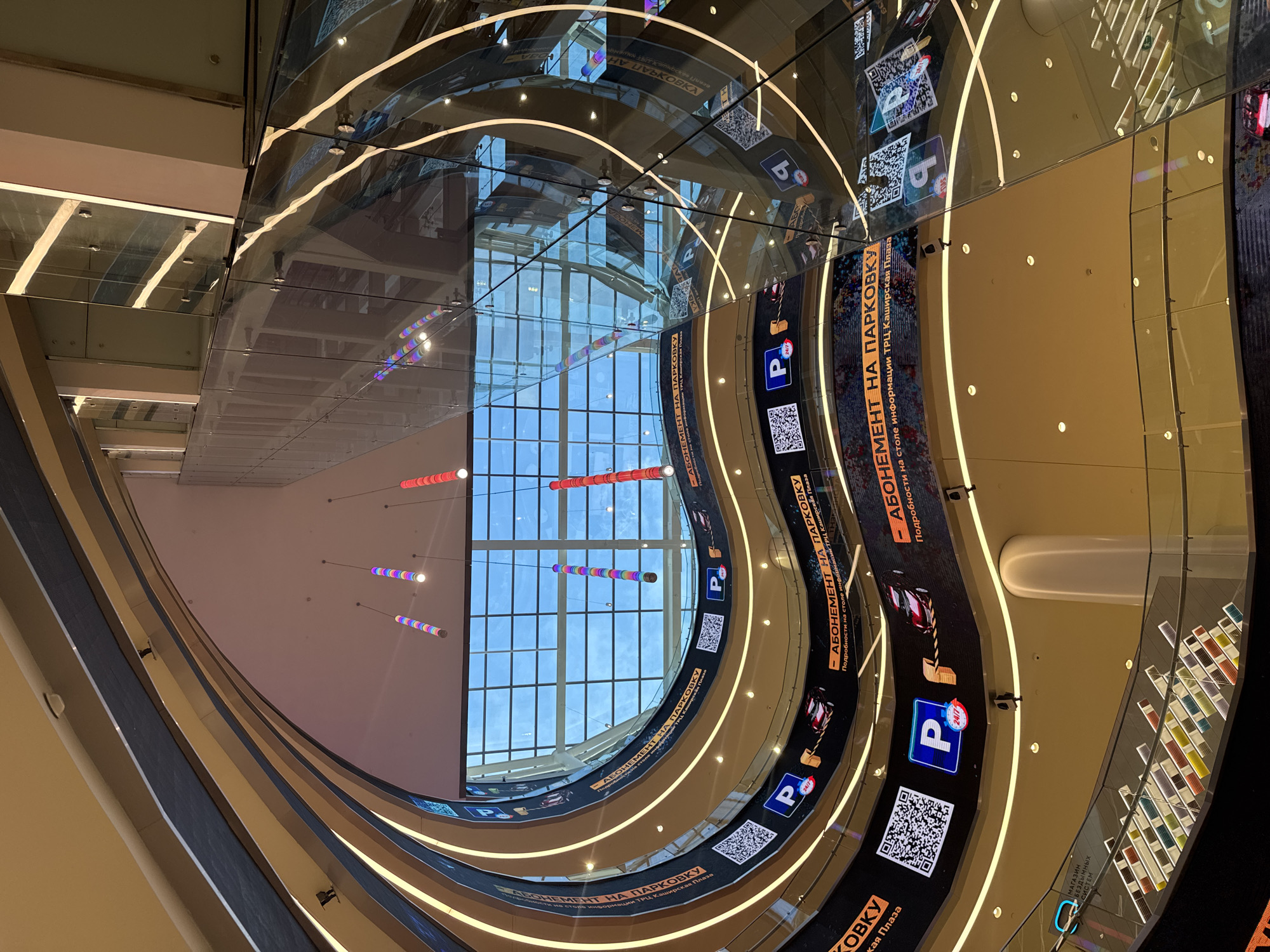 This is an interior shot of a multi-level shopping mall, likely the Kashirskaya Plaza shopping center, in Moscow, Russia. The image captures a unique architectural design featuring a large atrium with curved levels and a glass ceiling. The levels are lined with digital screens displaying advertisements, primarily for parking subscriptions. QR codes and parking symbols with "24/7" are prominently featured. Decorative, cylindrical lights in various colors hang from the ceiling. The overall impression is one of modern, dynamic commercial architecture. There are no people visible in the frame.