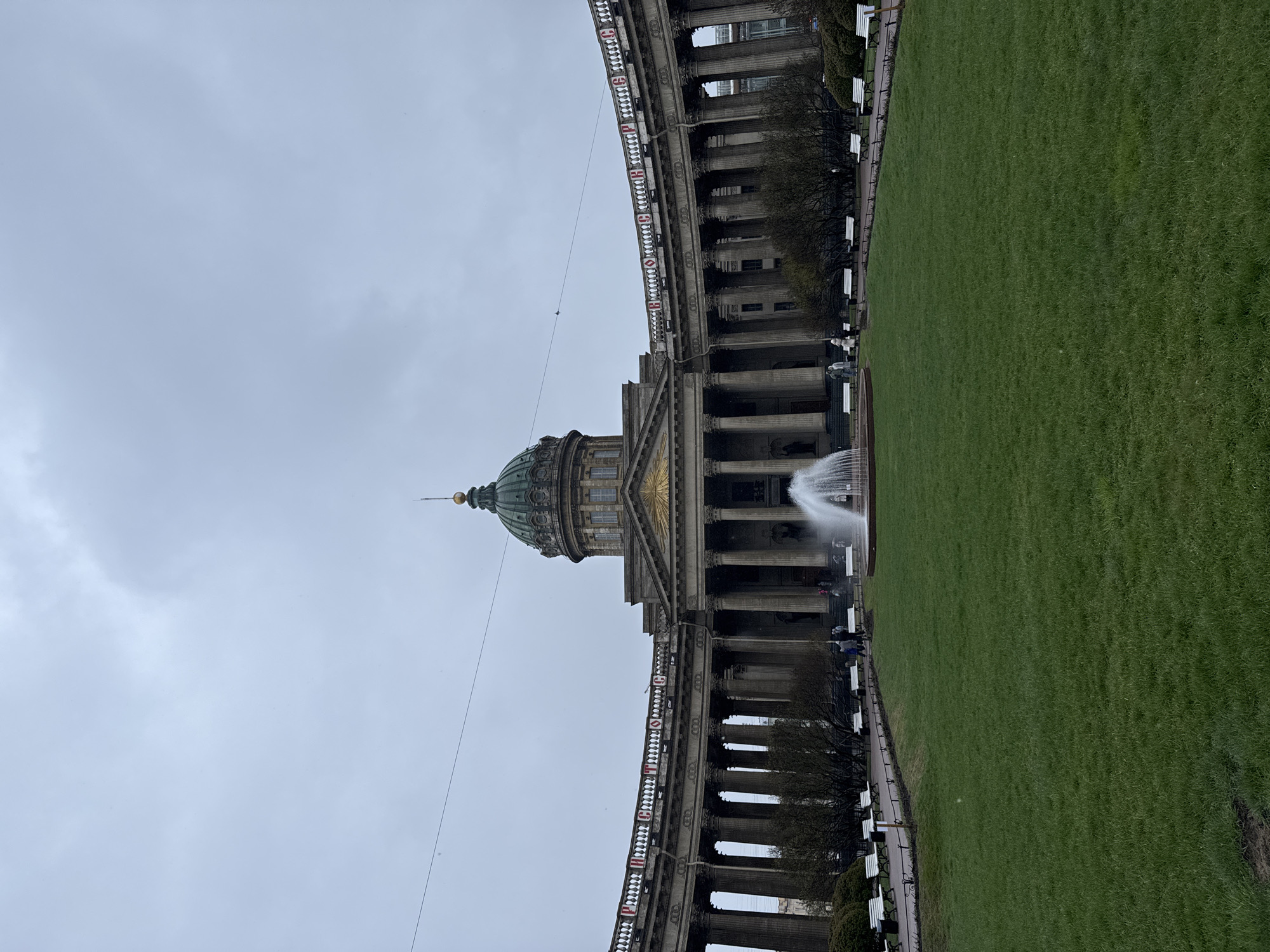 The image shows the Kazan Cathedral in Saint Petersburg, Russia. The cathedral, with its large dome and colonnades, faces onto a large green lawn. A fountain is in front of the cathedral. The sky is overcast. People can be seen walking around the fountain and sitting on benches along the lawn. There is a banner along the colonnade with Russian text.