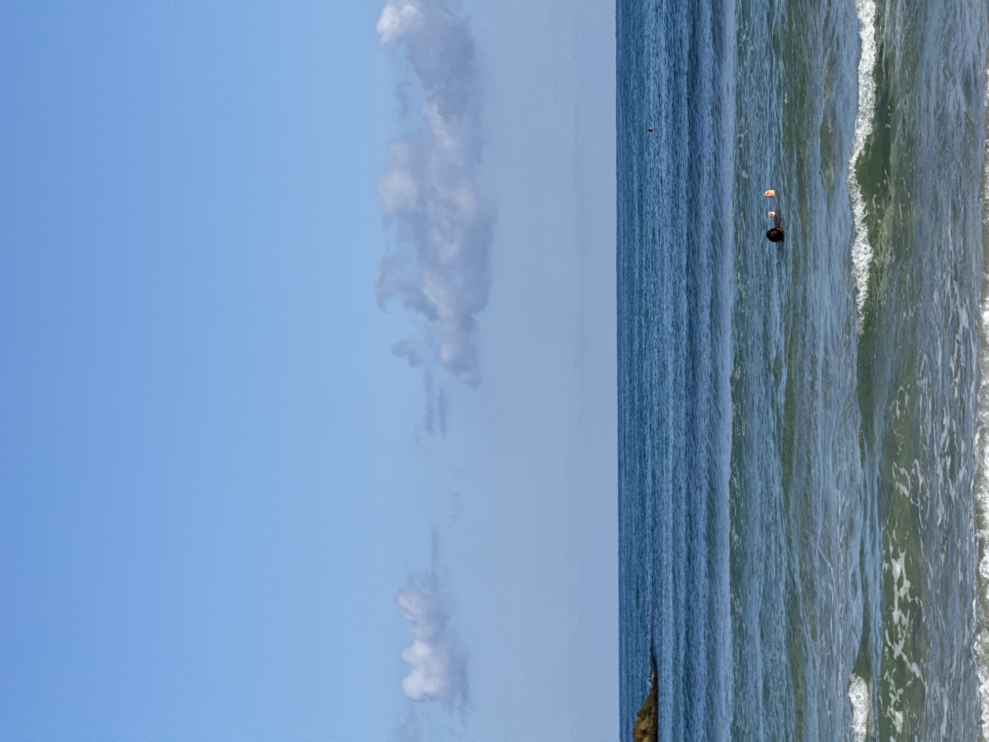 A person is floating on their back in the ocean, with their feet visible above the water. The person is wearing a dark garment. The ocean is calm with gentle waves and some white foam at the shore. In the distance, there is a rocky outcrop on the left side of the image and a clear blue sky with scattered white clouds. This scene is likely at a beach in Tel-Aviv, Israel, given the typical conditions and coastline.