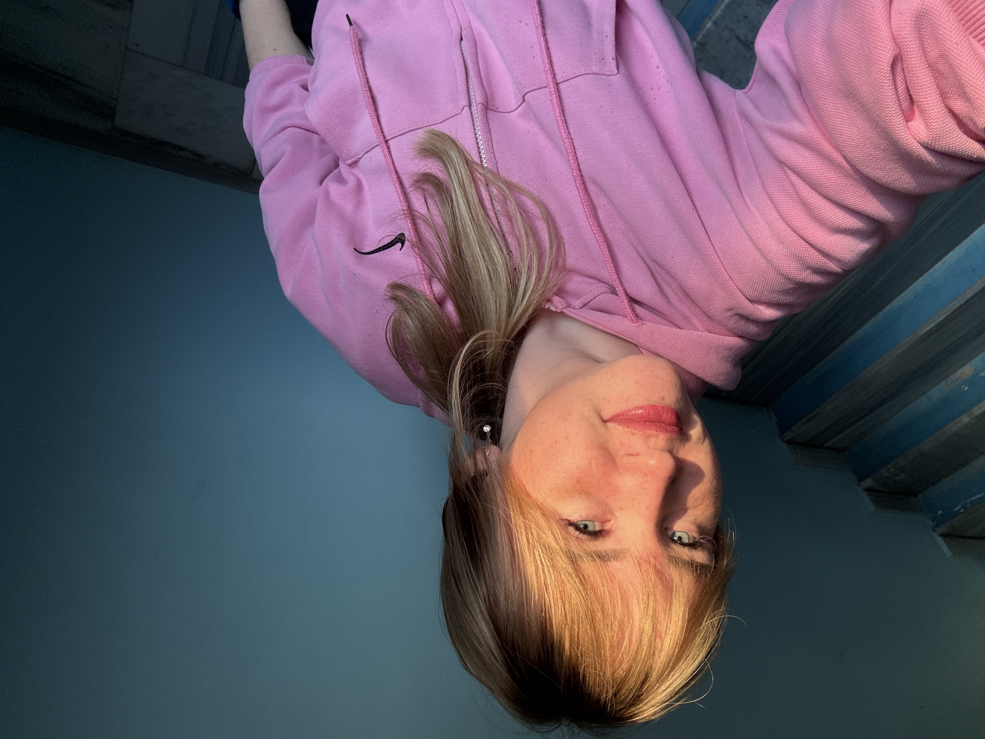 The image displays a close-up portrait of a young woman, rotated 90 degrees counter-clockwise. She has light-toned skin with visible freckles across her nose and cheeks, and bright, light-colored eyes (appearing blue or green). Her blonde hair is styled with bangs, framing her face, and falls loosely around her. She has pink or red lipstick on her lips and maintains a neutral to slightly smiling expression, looking directly at the camera. A small, silvery stud earring is visible in her right ear.
She is dressed in a bright pink hooded sweatshirt (hoodie) with a zipper down the front. A black Nike swoosh logo is clearly visible on the left chest area of the hoodie. The fabric of the garment shows a textured surface.
The background of the shot is divided. On the left side, there is a plain, dark teal or blue surface, resembling a wall. On the right side, a structured element with horizontal blue and gray stripes is visible, possibly depicting a wooden railing, steps, or paneling. The lighting is bright and focused on her face and hair, creating clear highlights, while the background remains in darker tones.
The activity taking place is a self-portrait or selfie. There are no explicit cues for the time of day or specific weather conditions, but the lighting suggests an indoor setting or a directed light source. The location context for this image is Марковский, Russia. The only visible text is the Nike logo on her hoodie.