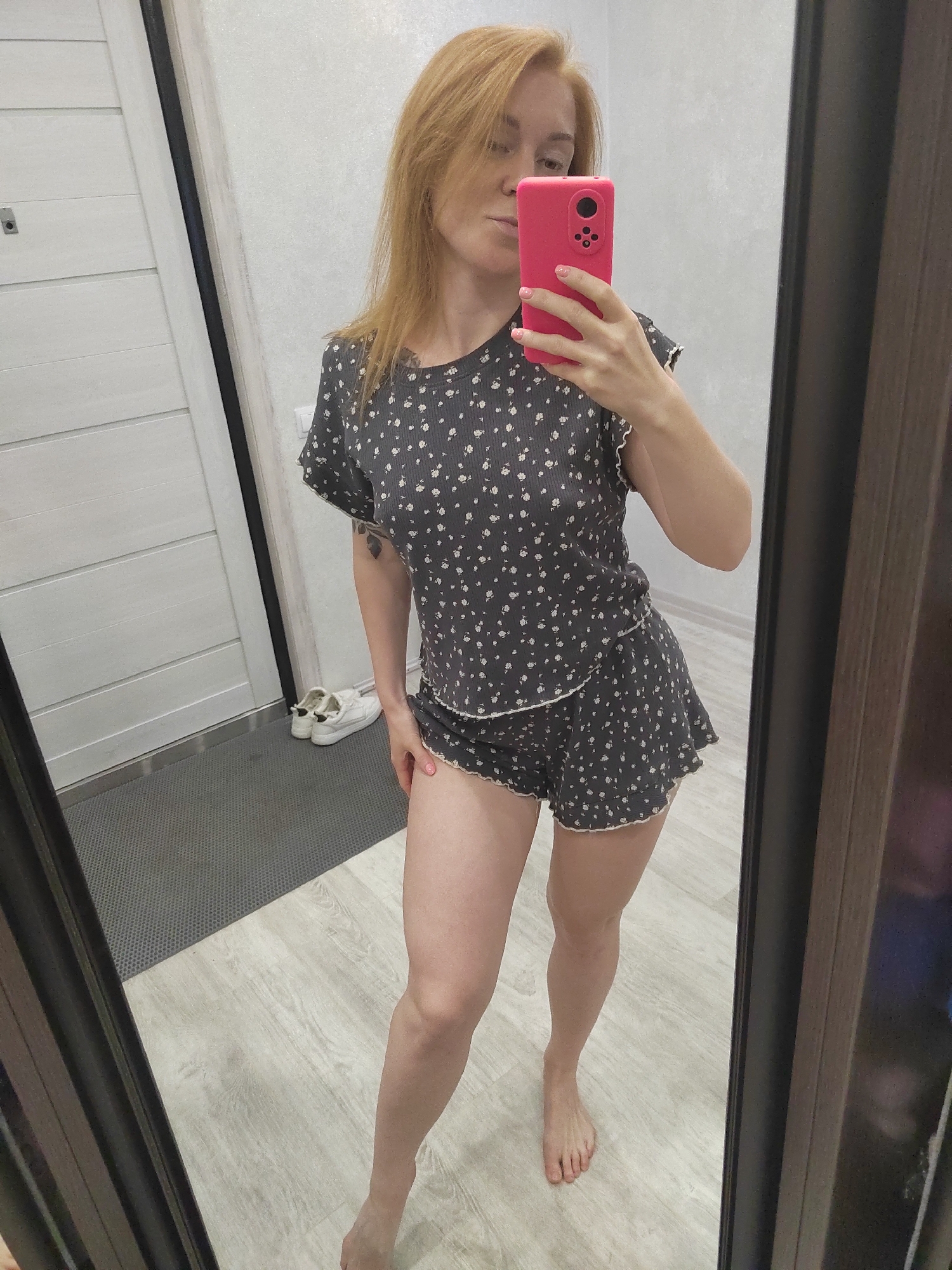 A woman with blonde hair stands in front of a mirror, taking a selfie with her pink smartphone. She is wearing a dark grey pajama set with a floral print and white trim. Her right arm is raised, holding the phone, and her left hand rests on her right thigh. She is barefoot. The background includes a white door with horizontal panels, a grey doormat, a pair of white sneakers, and a wooden floor. The mirror reflects the woman and the surrounding room. The scene appears to be an indoor domestic setting, possibly a hallway or bedroom, in an apartment in Saint Petersburg, Russia.