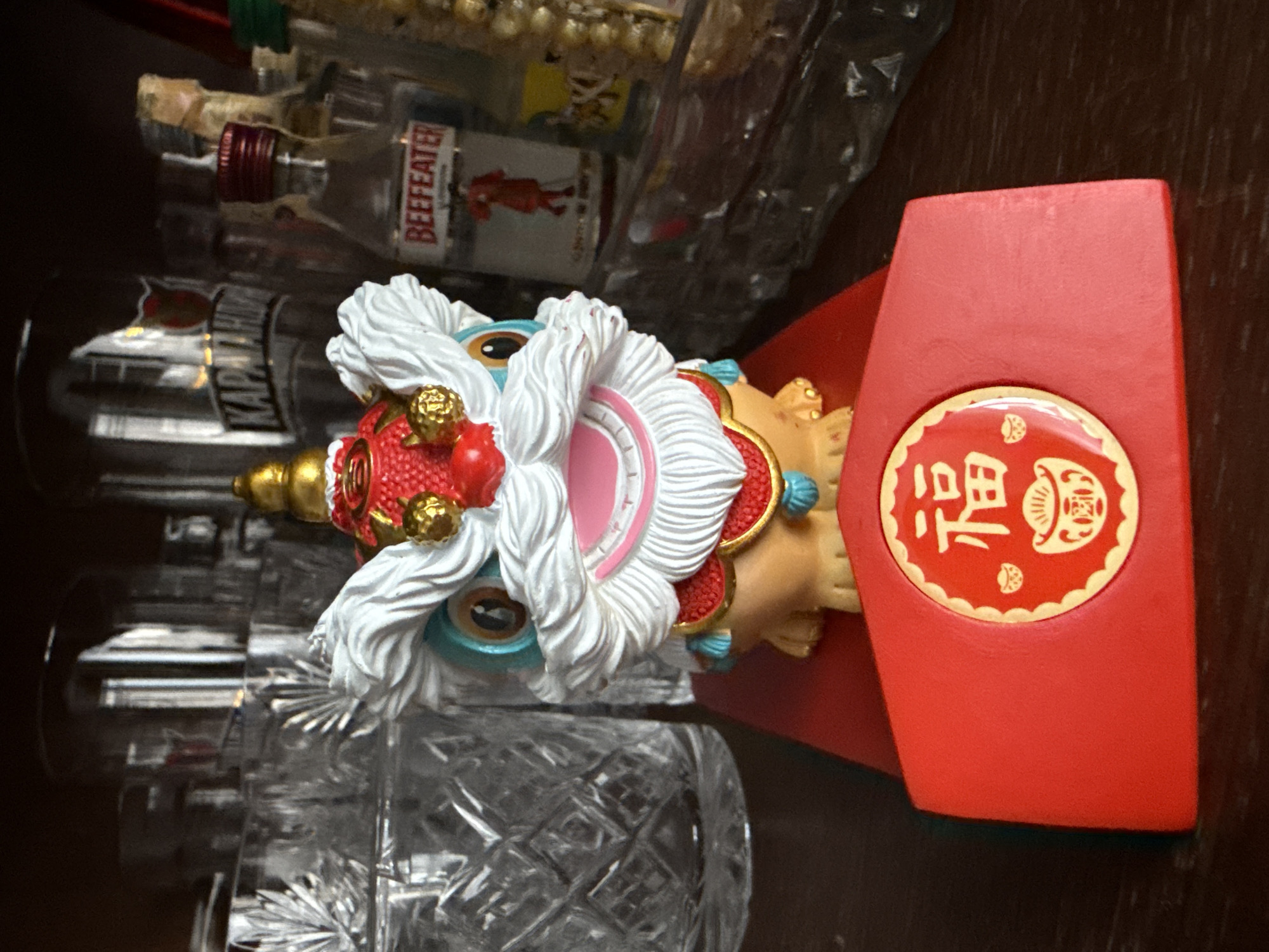A small, decorative lion statue sits on a red platform in front of several glass bottles on a dark shelf. The lion has a white mane and eyebrows, a red and gold headdress, and a tan body with red and blue accents. The red platform has a circular emblem with Chinese characters and a decorative border. The background shows a collection of various glass items, including glasses and bottles, some of which appear to be alcohol. One bottle in the background is labeled "BEEFEATER." The lighting is dim, casting shadows on the shelf. The image does not provide any information about the city of Tekeli, Kazakhstan.