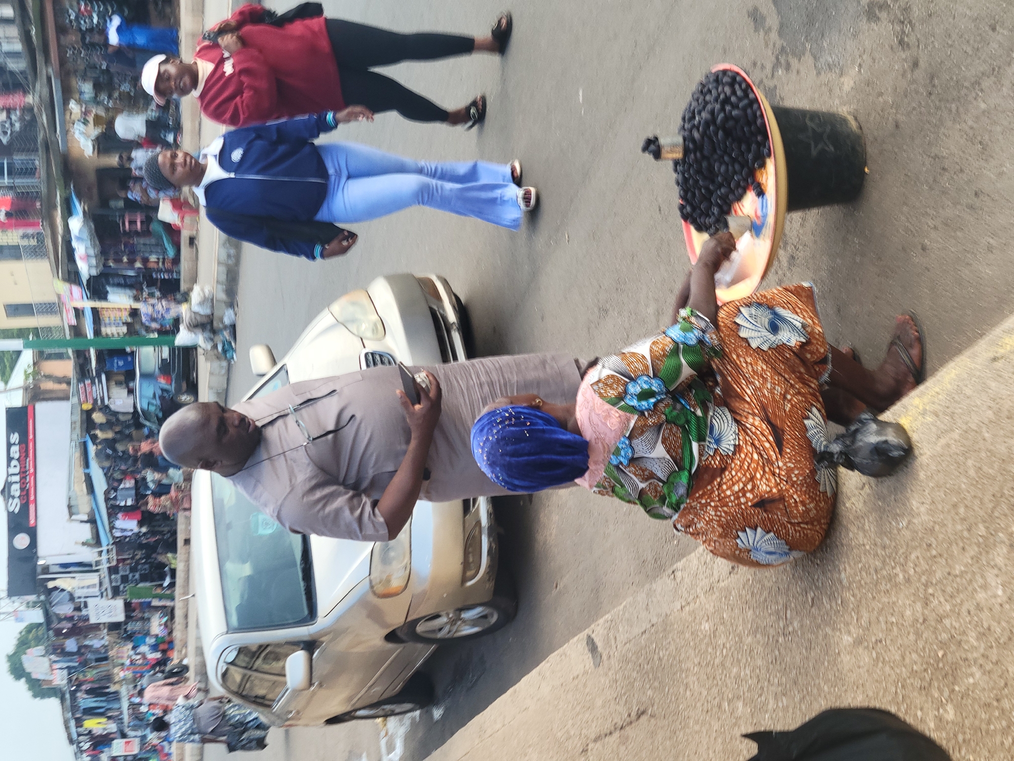 This image captures a street scene in Jos, Nigeria, where a vendor is selling a pile of dark-colored fruits, likely dates, from a colorful tray placed on top of a bucket. The vendor, a woman wearing a blue headscarf and a patterned orange and brown skirt, is seen reaching towards the fruit. In the background, a gold-colored sedan is parked on the road, and other pedestrians and market stalls are visible, suggesting a bustling urban environment. A man in a light brown shirt is standing near the car, facing away from the camera. Two other women are walking in the background, one in a blue jacket and light blue pants, and the other in a red sweatshirt and dark pants. The ground appears to be paved, with a concrete curb in the foreground.