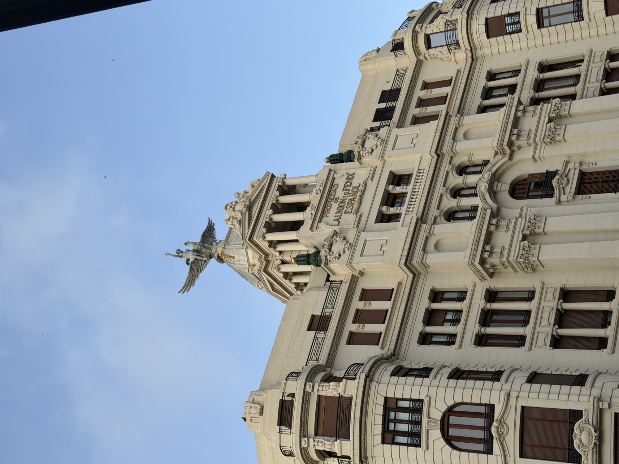The image shows the facade of a light-colored building with classical architectural elements and a statue on top. The building has multiple stories with numerous windows, some of which are arched. Balconies with black railings are visible on some floors. The building's facade is adorned with decorative carvings and columns. On the rooftop, there is a prominent sculpture of a winged figure, possibly an angel or a mythological being, mounted on a pedestal. Above the sculpture, the sky is a clear blue with a few small dark birds in flight. The text "PROPIEDAD DE LA UNION Y EL FENIX ESPAÑOL" is inscribed on the facade.