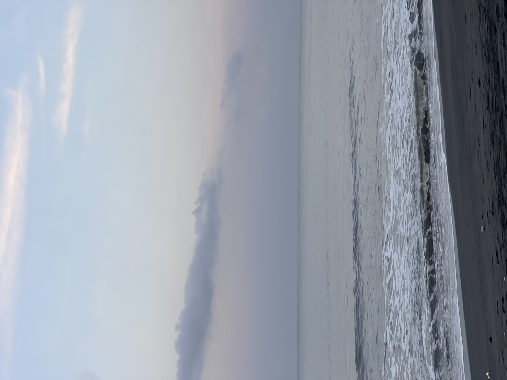 The image depicts a coastline. A grayish sky occupies the left side, with some visible clouds. The ocean extends to the right, showing waves crashing onto a dark beach. The picture's orientation is rotated ninety degrees counter-clockwise. Given the location of the city of Kediri, Indonesia, the location is likely a beach in the Southern coast of Java or on one of the nearby smaller islands in Indonesia. There are no people visible in the scene.