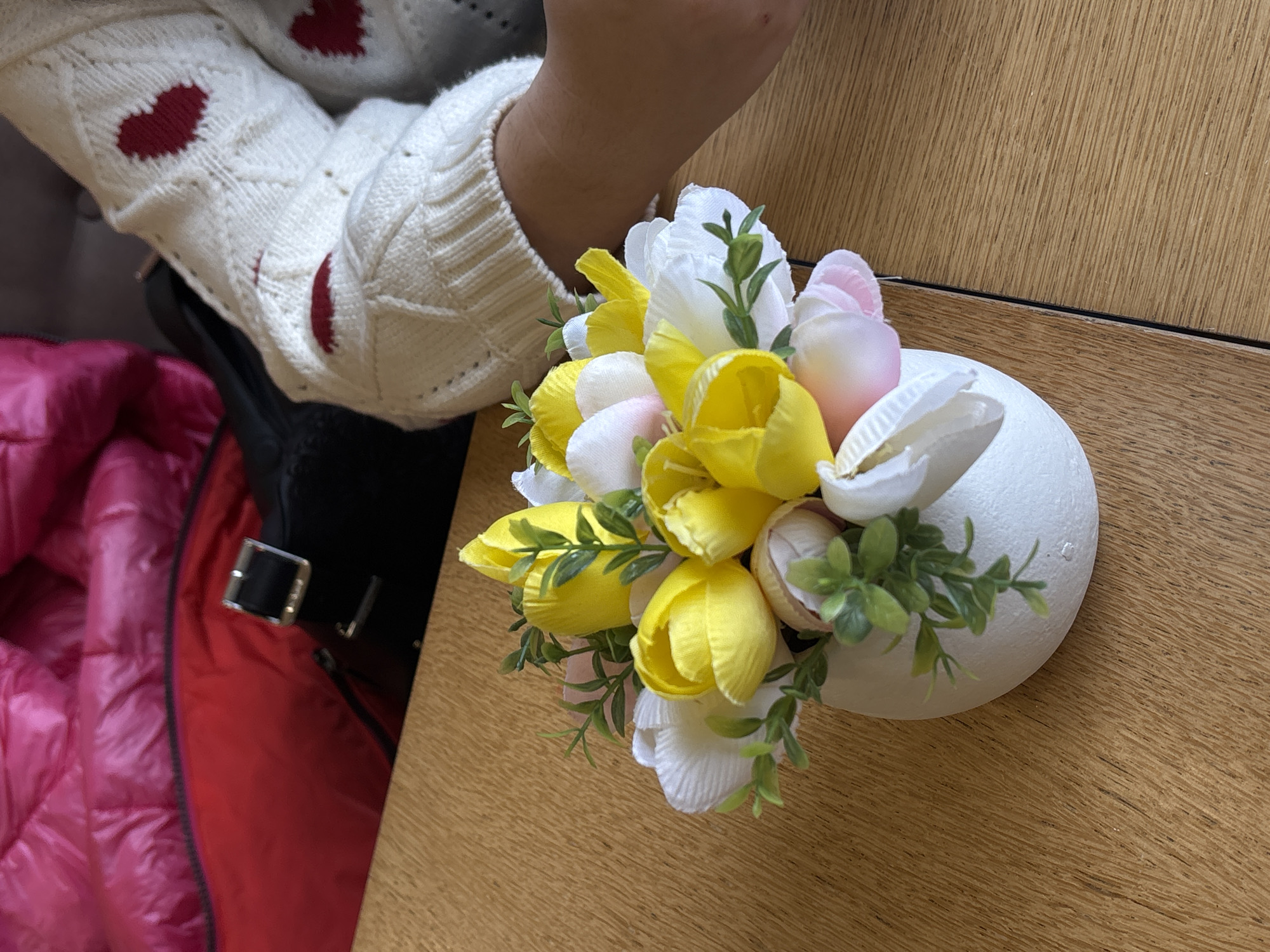 A top-down shot captures a floral arrangement in a white egg-shaped vase resting on a wooden surface, likely a table, in Varna, Bulgaria. The arrangement is composed of yellow, white, and pink tulips interspersed with green foliage. Part of a person's left arm and the shoulder of a white knit sweater with red heart patterns are visible. Adjacent to these elements is a red, possibly padded, coat or jacket with a visible black buckle. The background suggests an indoor environment, likely a home or cafe.