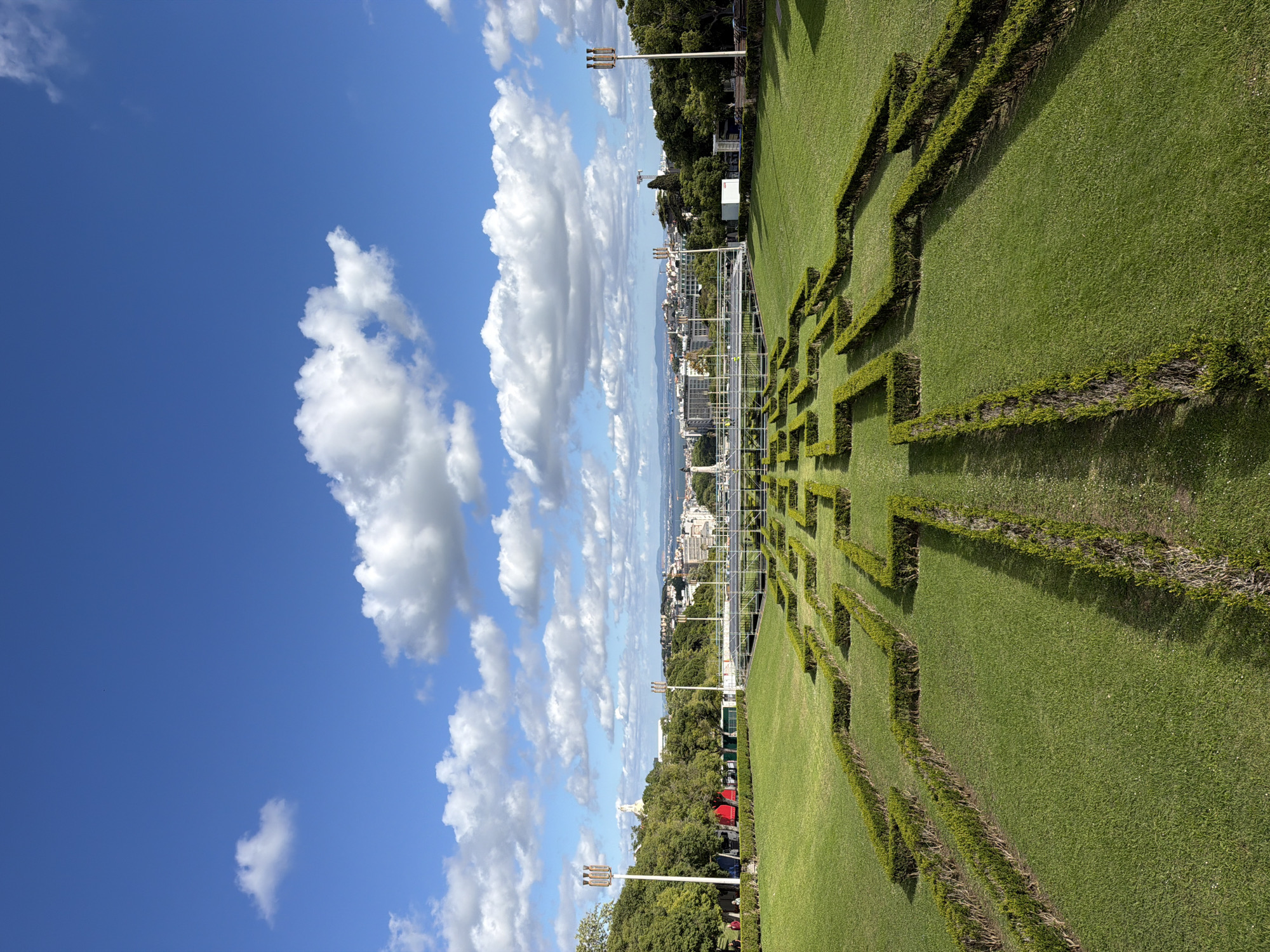 The image depicts a bright, sunny day in Lisbon, Portugal, specifically in Eduardo VII Park. The sky is a vibrant blue with scattered cumulus clouds. The park features a geometric hedge maze, neatly maintained, with pathways of closely-cut grass. In the distance, the city of Lisbon can be seen with its characteristic architecture. A metal scaffolding structure rises within the park, likely for an event or exhibition. Streetlights are visible along the park's edge, suggesting a public space. There is the presence of trees and a lawn. No people are clearly visible in the image.