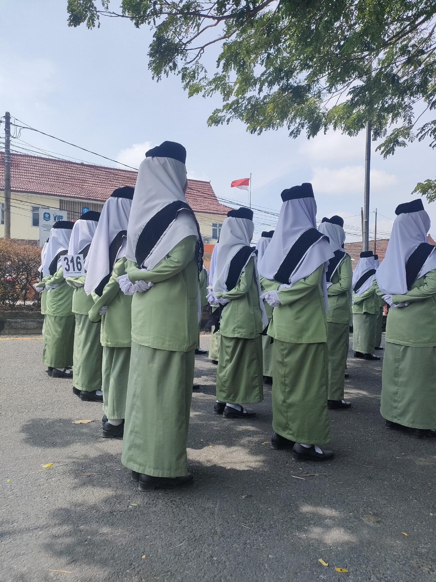 A group of young women in matching light green uniforms and white hijabs with black trim stand in a line on a paved road. They are facing away from the camera, with their hands clasped behind their backs. One woman in the front row has a white sign with "316" printed in black on it attached to her uniform. In the background, there are buildings with red tiled roofs and a flagpole with a red and white Indonesian flag. Trees with green leaves are visible in the upper part of the image. The scene appears to be outdoors, possibly in a school or public area, under a bright sky. The location is the city of Sungailiat, Indonesia.