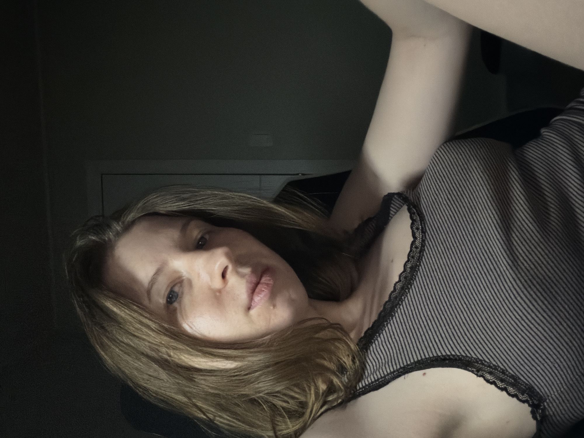 The image is a close-up, low-angle, indoor photograph of a young woman with shoulder-length light brown hair. She is wearing a black and grey striped tank top with black lace trim. Her left arm is extended forward and out of frame. The background is dark and indistinct, appearing to be an interior room with a dark wall and a sliver of a doorway visible on the right. The lighting is dim, casting shadows and highlighting the woman's face and upper body. The photograph does not contain any discernible landmarks or features that would indicate its location in Dongducheon-si, South Korea.