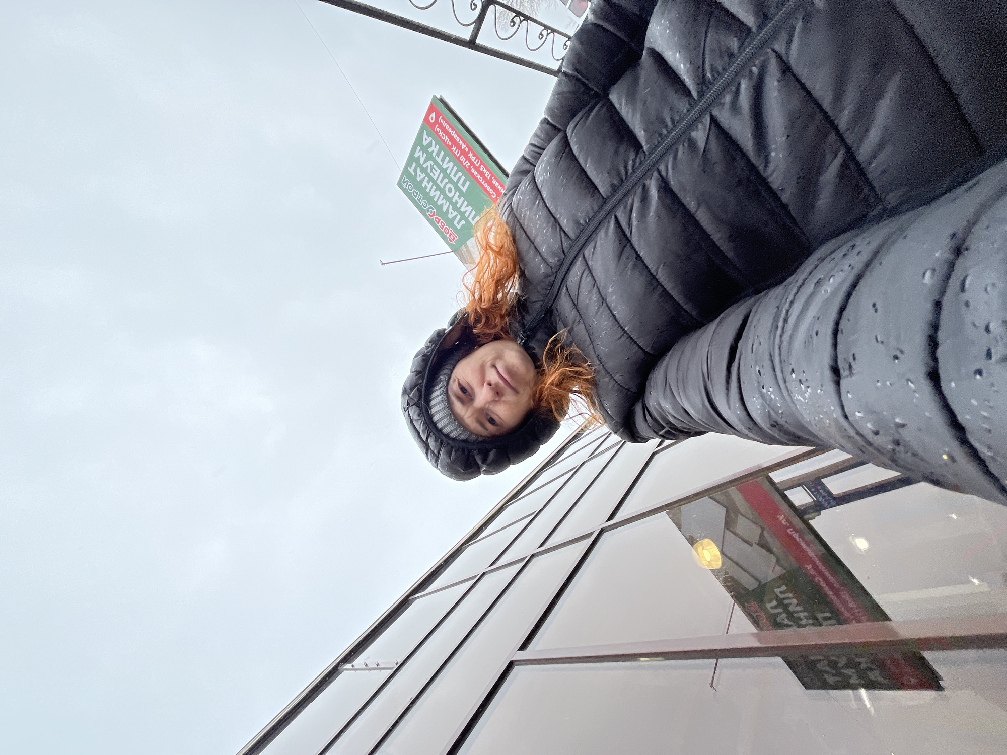 An upside-down selfie captures a woman with red hair looking directly at the camera, wearing a black puffer jacket covered in small water droplets, and a gray-striped beanie. The image is taken from a low angle, pointing upwards towards an overcast, light gray sky.
The woman's jacket suggests rainy or wet weather. Behind and above her head, an informational sign is visible, also upside down from her perspective. The sign is green with white text, listing types of building materials and store locations.
Visible text on the sign reads:
- "ДОБРОСТРОЙ" (DOBROSTROY)
- "ЛАМИНАТ" (LAMINATE)
- "ЛИНОЛЕУМ" (LINOLEUM)
- "ПЛИТКА" (TILES)
- Two addresses for what appears to be store branches:
- "Советская, 2/10 (ТК «ЦСК»)" (Sovetskaya, 2/10 (TC "TsSK"))
- "Сенная, 13к5 (ТРК «Аквареал»)" (Sennaya, 13k5 (TRK "Akvaryel"))
Below the woman, a modern building with large, reflective glass windows is visible, showing reflections of the sky and the lower part of the sign. The overall scene suggests an urban setting in Chaikovsky, Russia, during daytime under cloudy or drizzly conditions. The unique perspective makes it appear as if the woman is hanging upside down from the building.