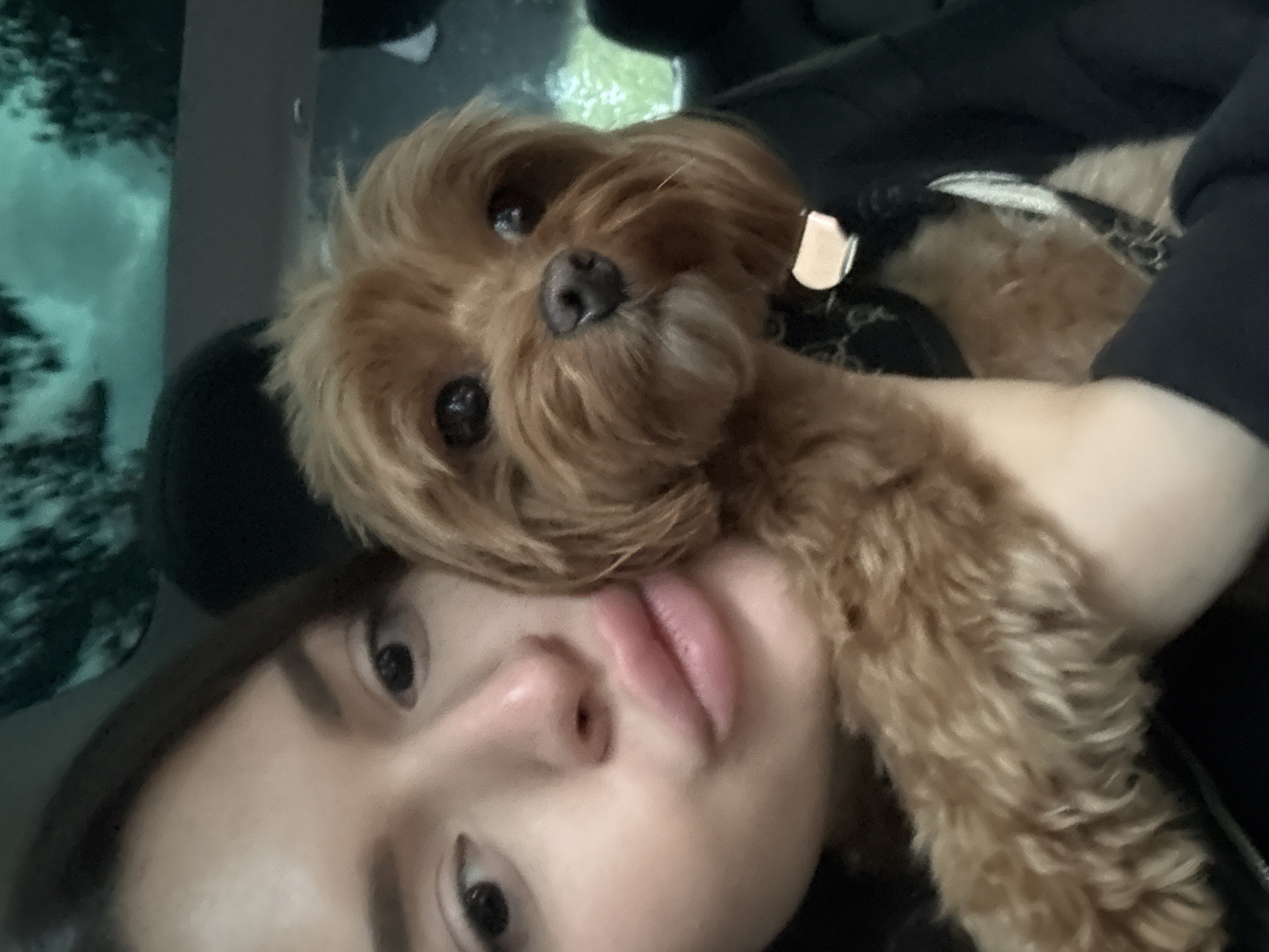 A close-up, selfie-style photo shows a woman and a small, brown, fluffy dog inside a vehicle. The woman is in the foreground, her face close to the camera, with her lips pursed. She has brown hair and is wearing a dark-colored top. The dog, positioned next to her, is also looking towards the camera with a dark nose and eyes. The background is blurred and appears to be the interior of a car, with a dark seat and a hint of green foliage and sky visible through a window. The overall impression is an informal snapshot taken during a car ride. There is no indication that this photo was taken in Prague, Czechia.