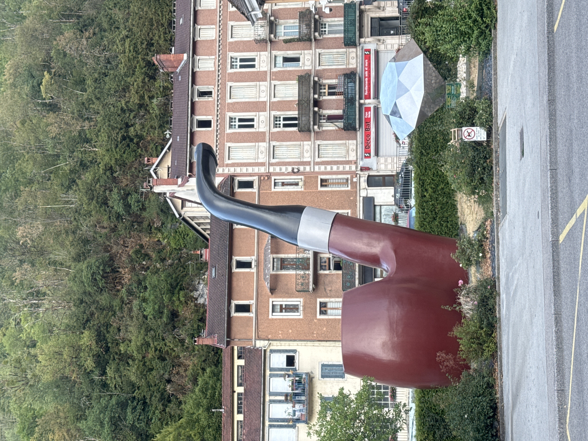 In Saint-Claude, France, a large, red, pipe-shaped sculpture stands prominently in front of buildings on a street. To the right of the pipe sculpture, a large, faceted, diamond-shaped sculpture is also visible. The background features a dense, green hillside covered with trees. The street in the foreground has a paved surface with yellow lines. The buildings appear to be multi-story structures with balconies and windows. The scene depicts an outdoor public space with prominent art installations.